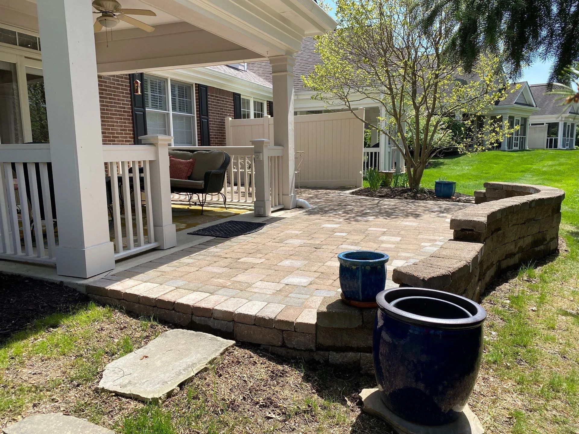 Stone patio beside a white porch with two blue pots and a raised retaining wall in a sunny backyard