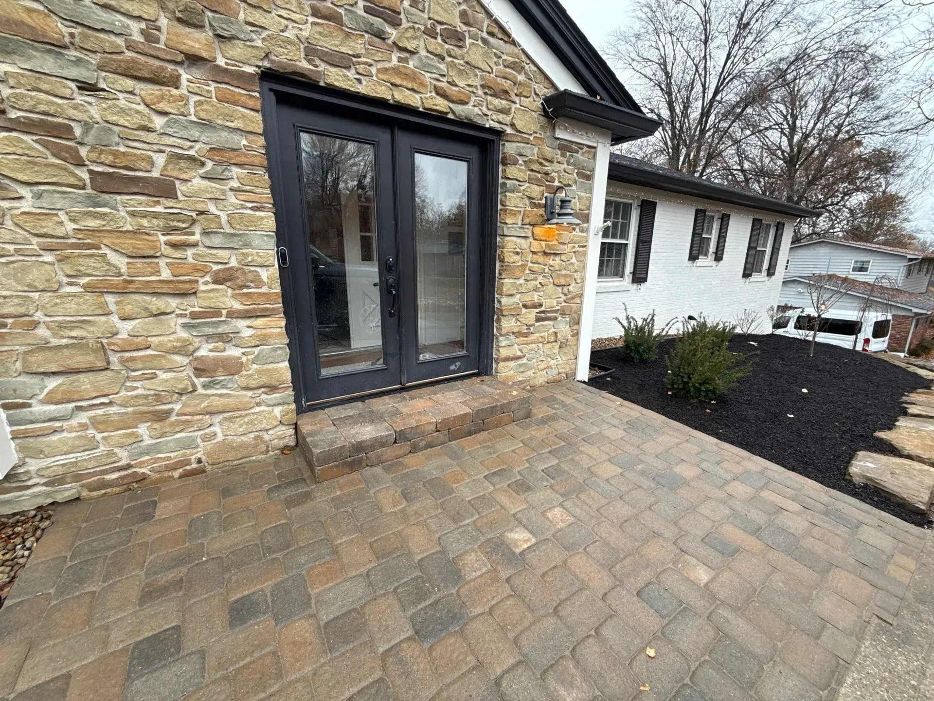 Stone patio with paver walkway beside a house, leading to black double doors and a white building nearby