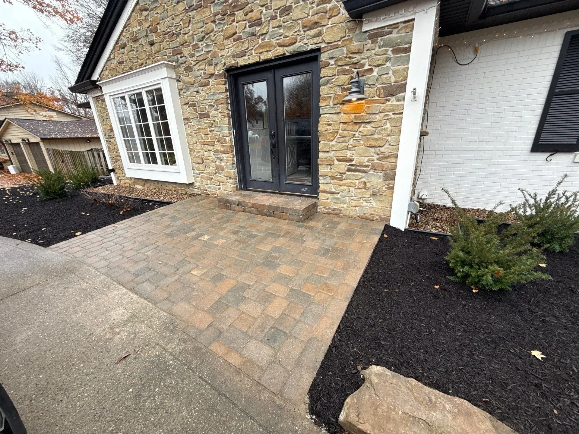 Stone house entry with black double doors, white-framed window, and paved driveway beside dark landscaping
