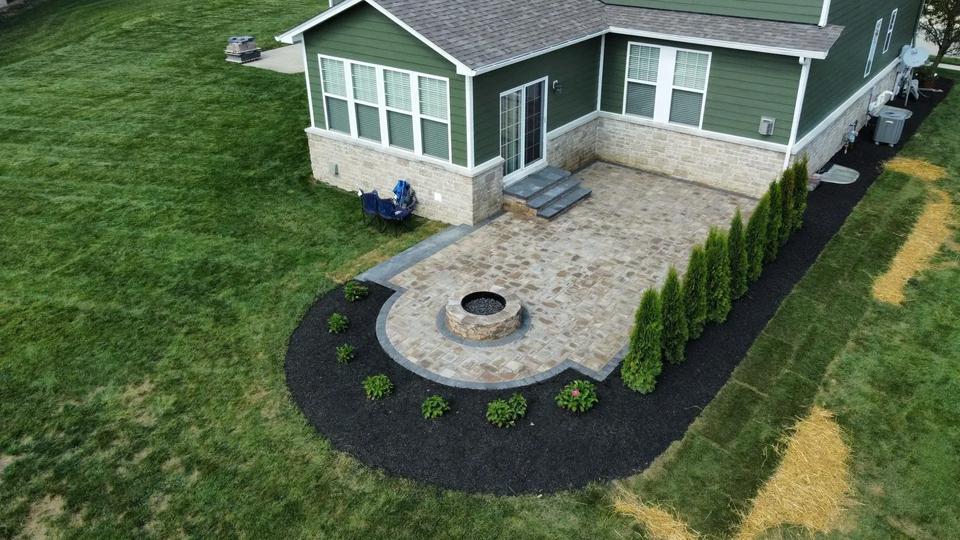 House with curved stone patio, fire pit, and landscaped front yard with shrubs and black mulch.
