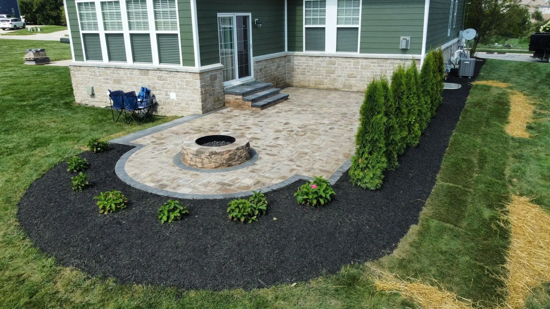 Backyard patio with stone fire pit, curved paver walkway, and landscaped shrubs beside a house