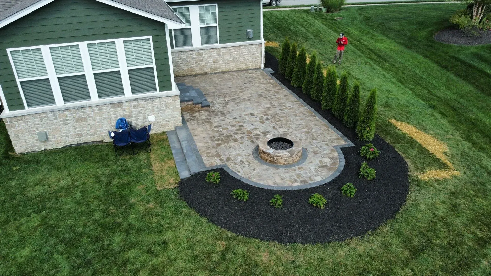 Stone patio with fire pit beside a house, bordered by mulch and shrubs, viewed from above