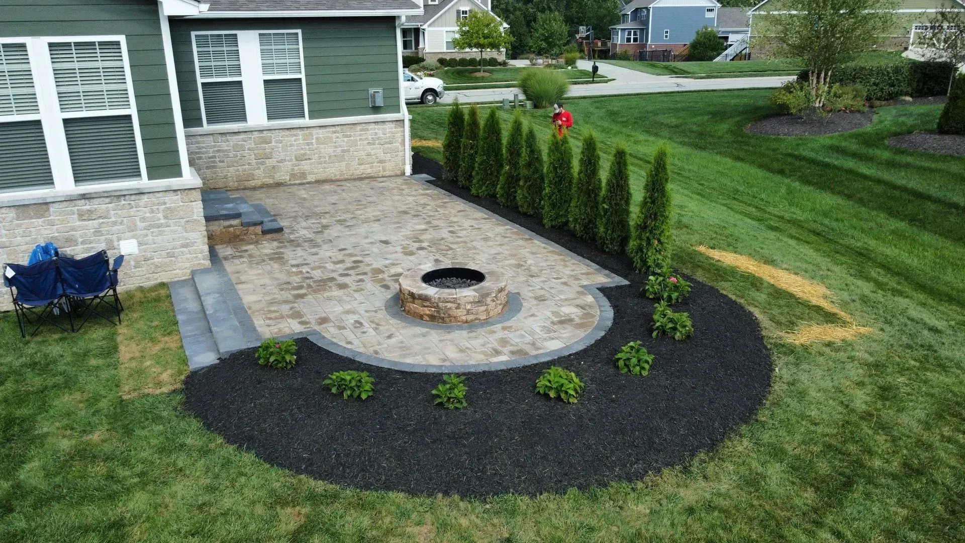 Patio with circular fire pit, stone pavers, and black mulch border beside a house and lawn