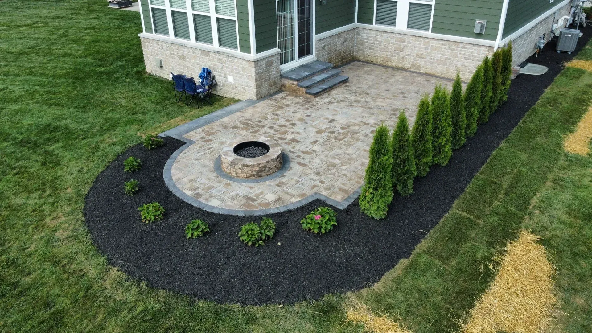 Curved stone patio with circular fire pit, bordered by mulch and shrubs beside a house lawn