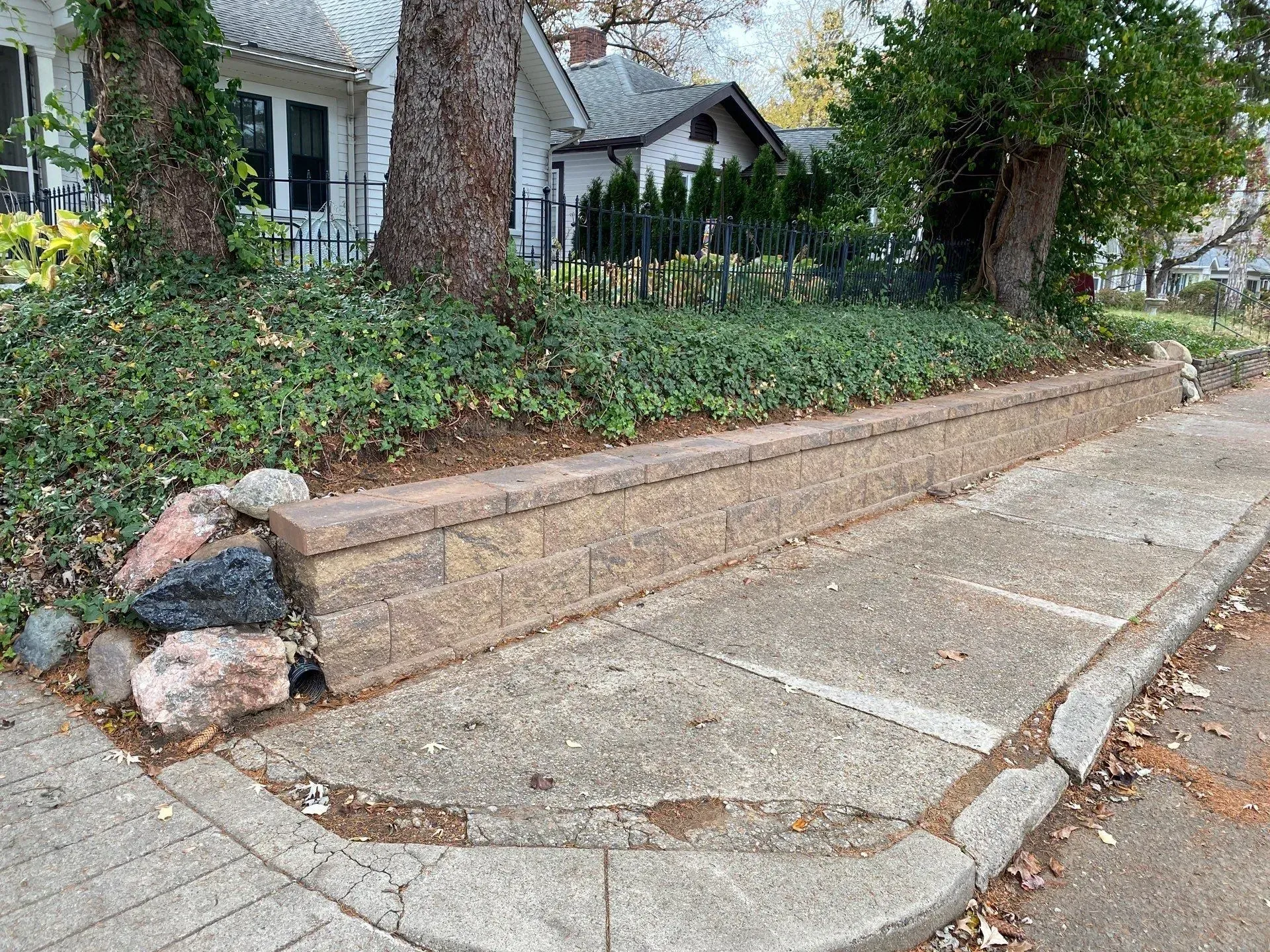 Concrete sidewalk with a raised stone planter retaining wall beside a house and trees