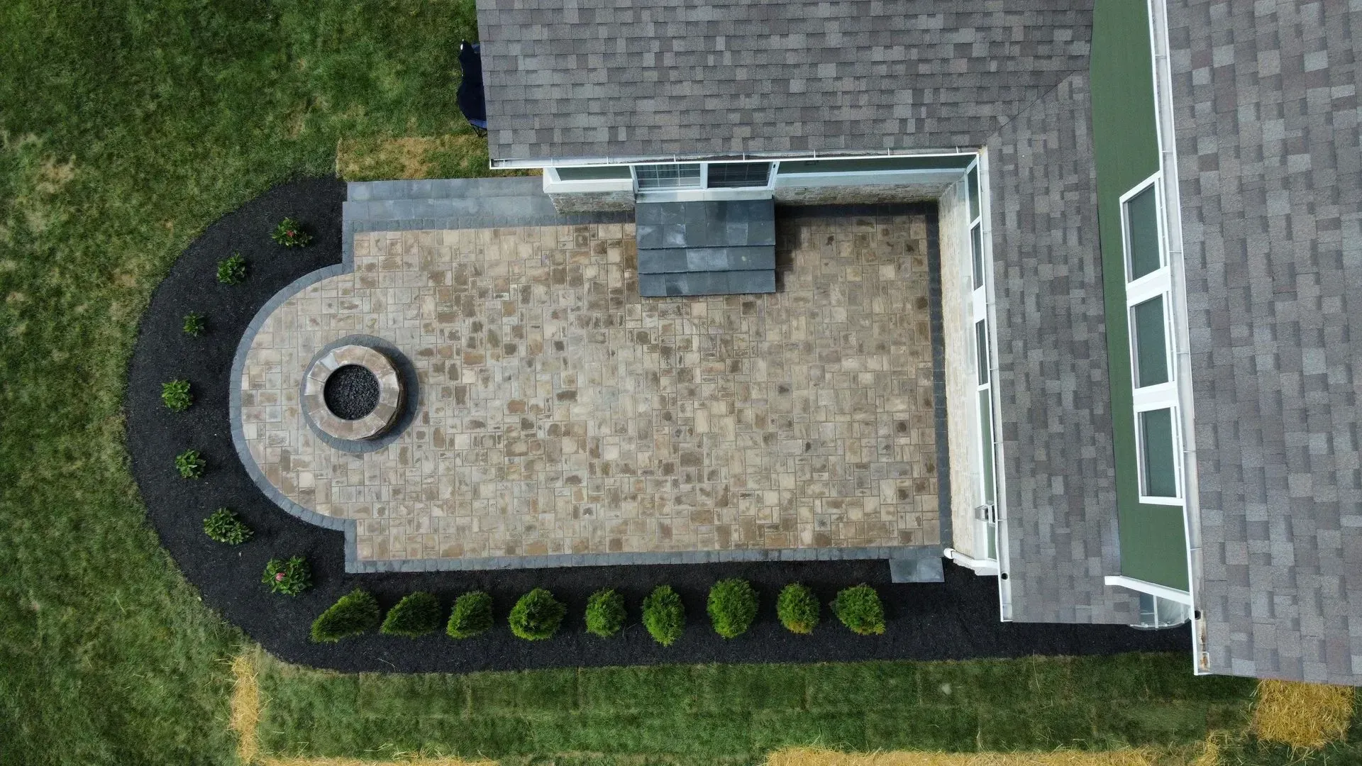 Aerial view of a backyard patio with stone pavers, circular fire pit, shrubs, and house roofline.