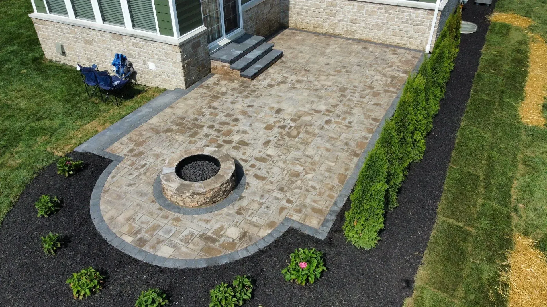 Stone patio with round fire pit beside a house, bordered by dark mulch and trimmed shrubs.
