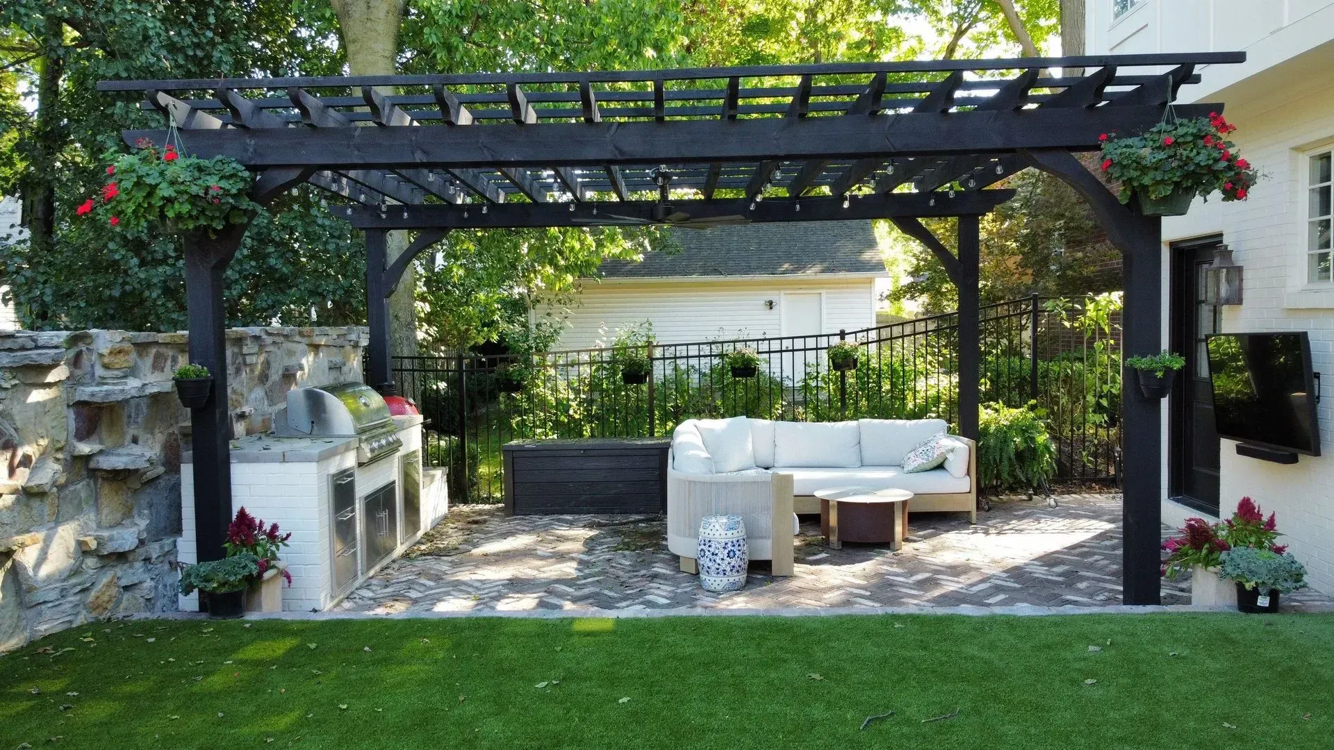Outdoor patio with dark pergola, white sectional sofa, and stone grill under lush greenery