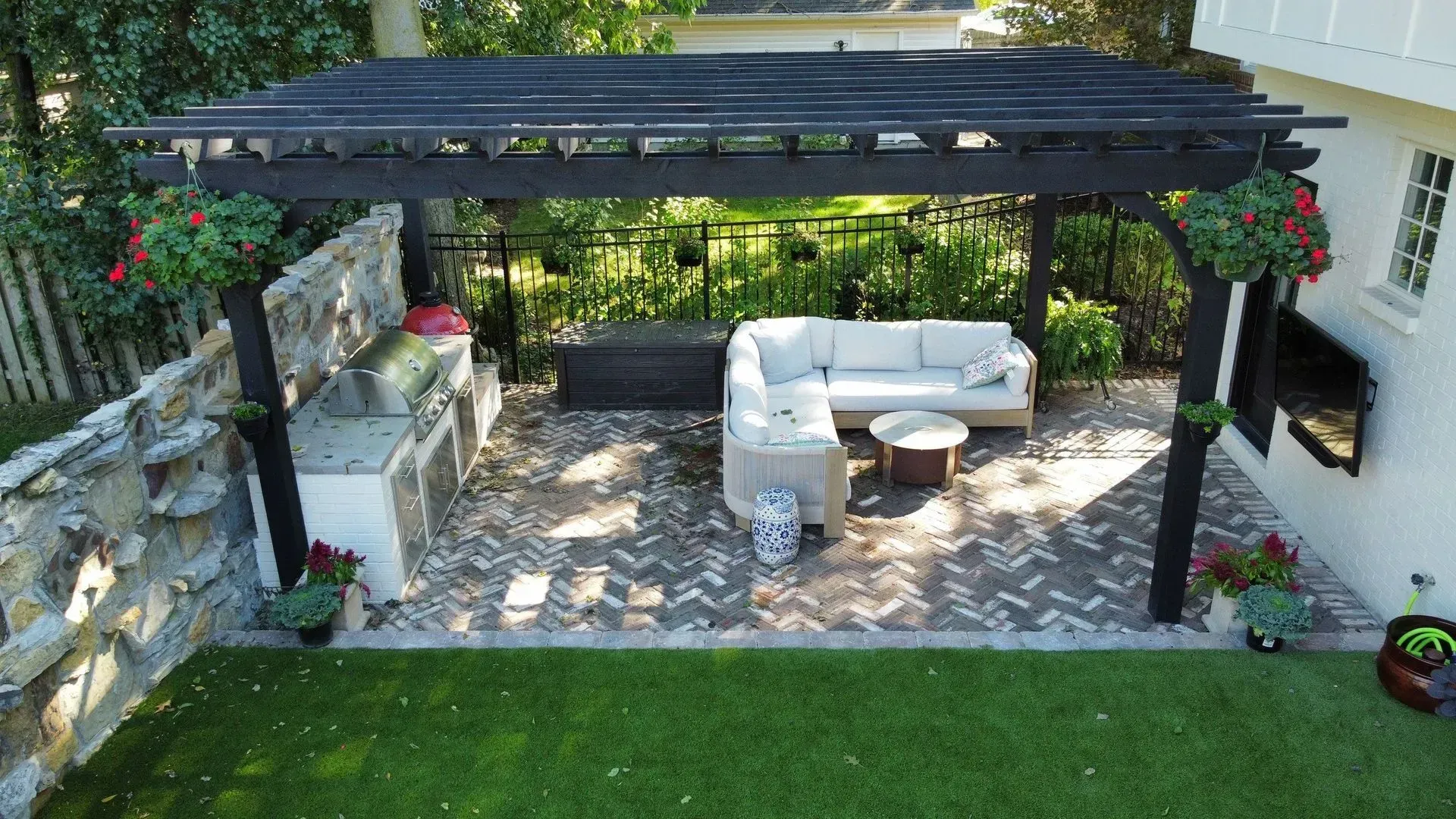Outdoor patio with pergola, white sofa, hanging flower baskets, and green lawn beside stone wall