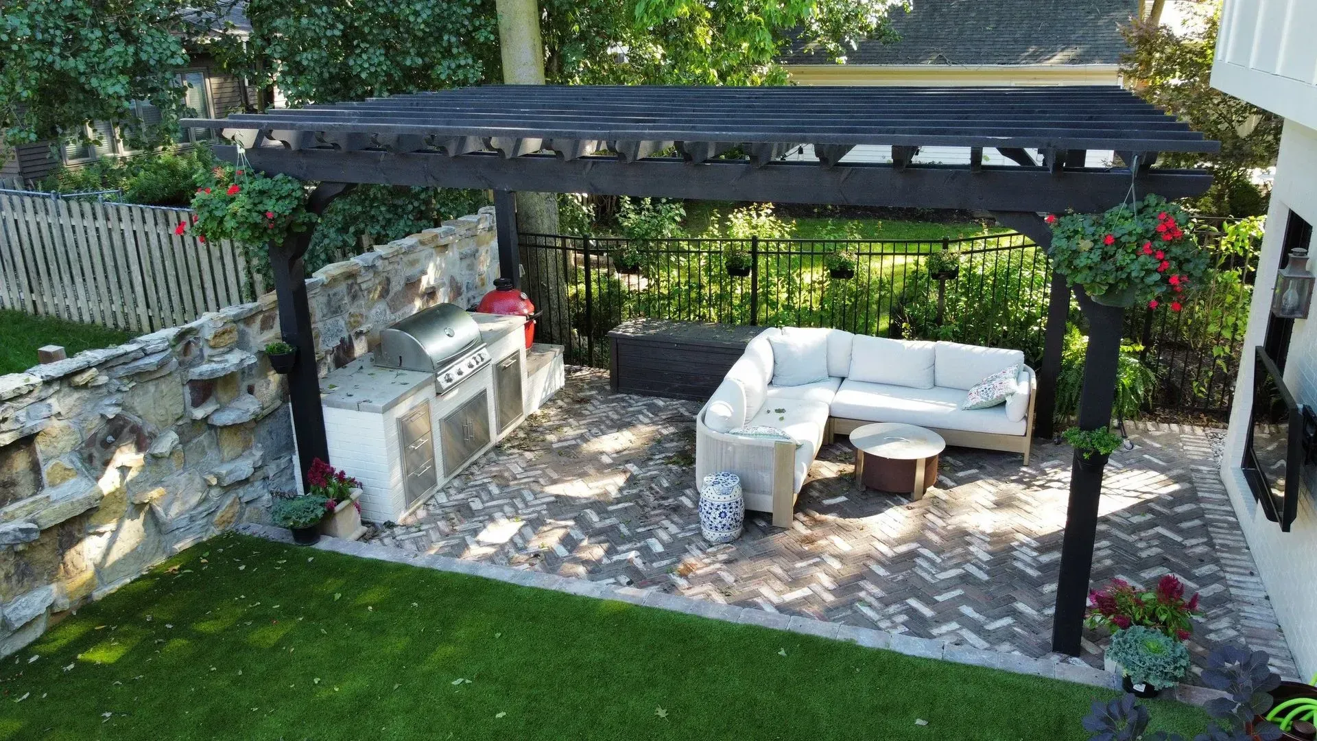 Covered patio with white outdoor seating, built-in grill, and stone paving beside a green lawn