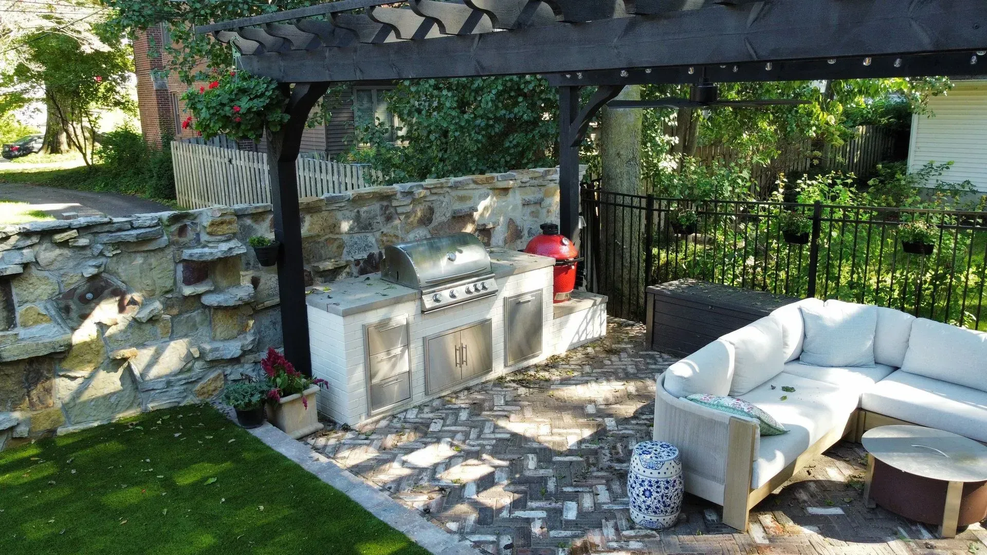 Outdoor stone patio with white sectional sofa, fire pit, and black pergola beside a garden wall
