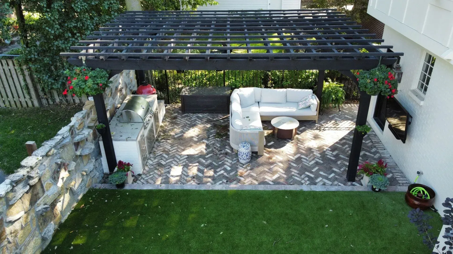 Stone patio with pergola, white outdoor seating, hanging flowers, and a green lawn beyond.