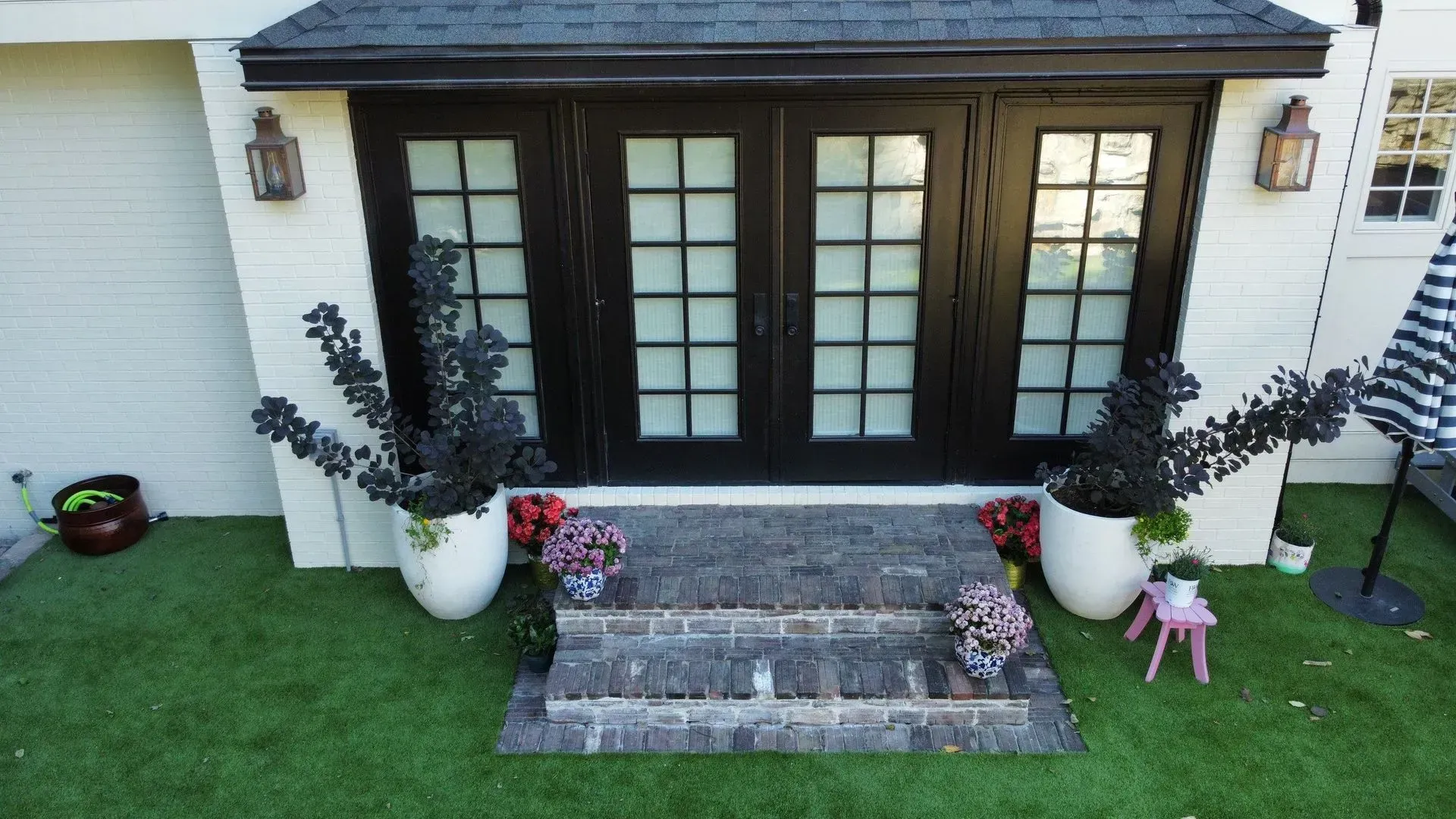 Front porch with black French doors, potted plants, and stone steps leading to a green lawn.