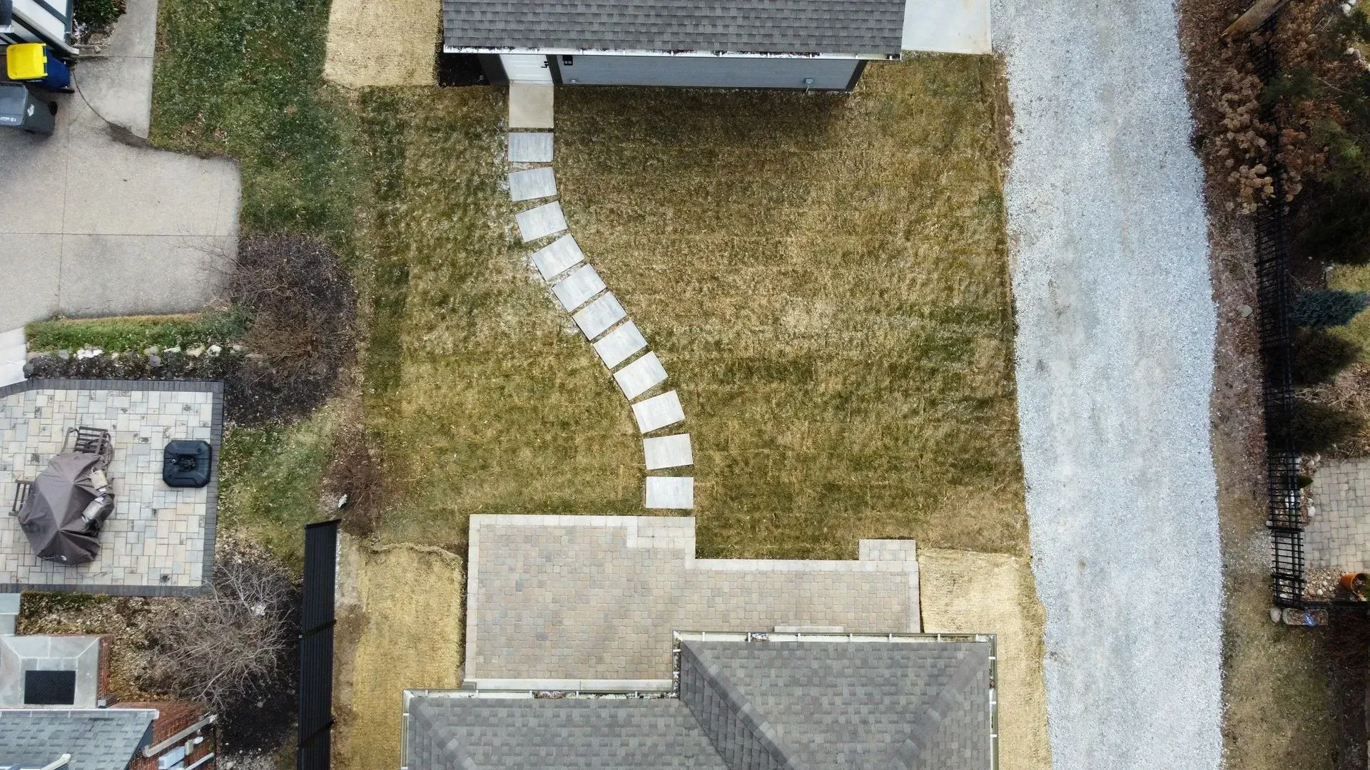 Aerial view of a backyard with a curved stone path, lawn, patio, and surrounding fences and houses.