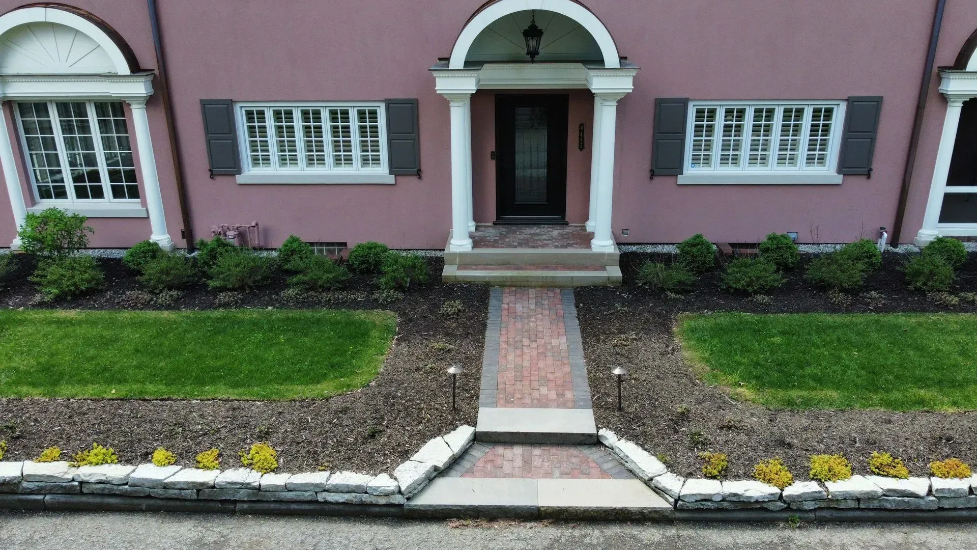 Front entrance of a pink house with a brick walkway, white-trimmed windows, and landscaped flower beds.