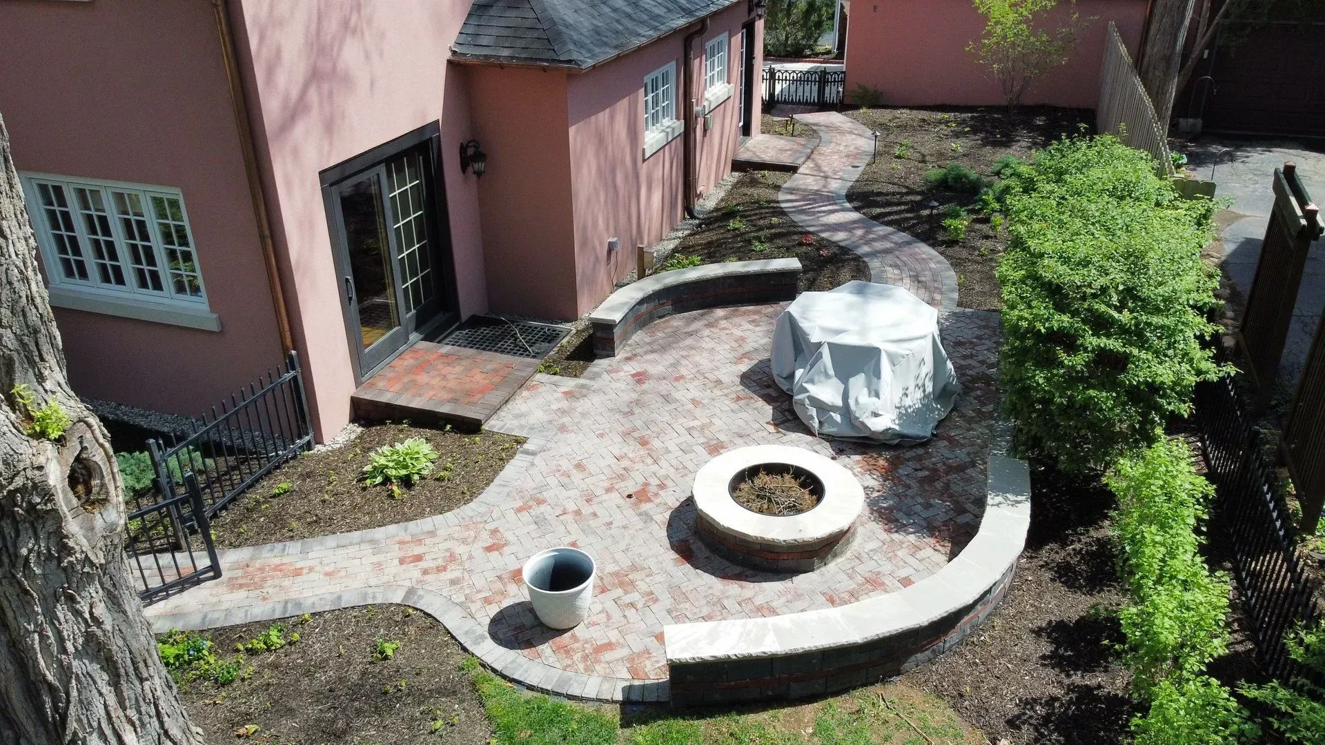 Curved garden patio beside a pink house, with a circular fire pit and white outdoor table under sunlight