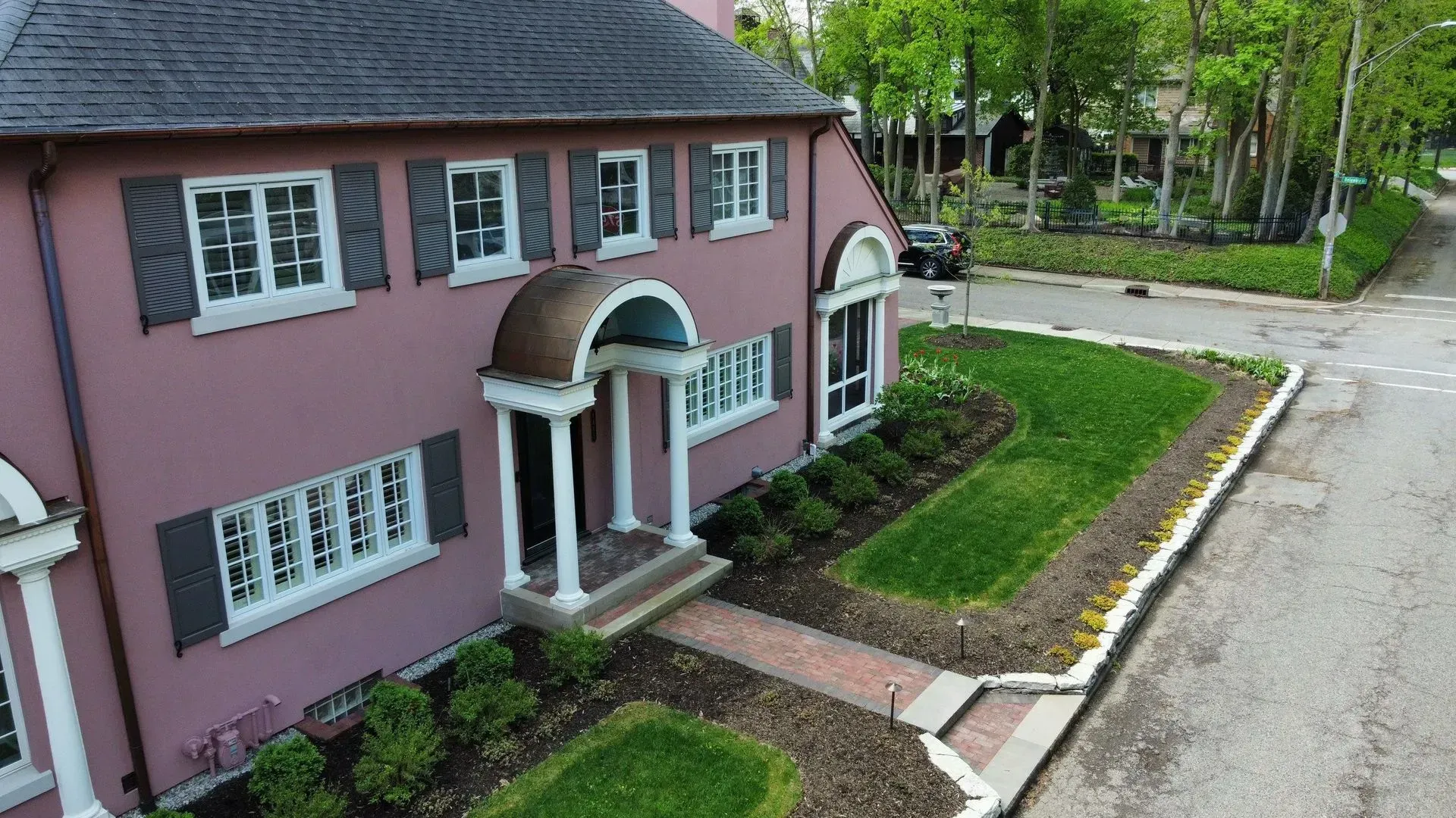 Pink two-story house with white trim and a front porch beside a road and garden.