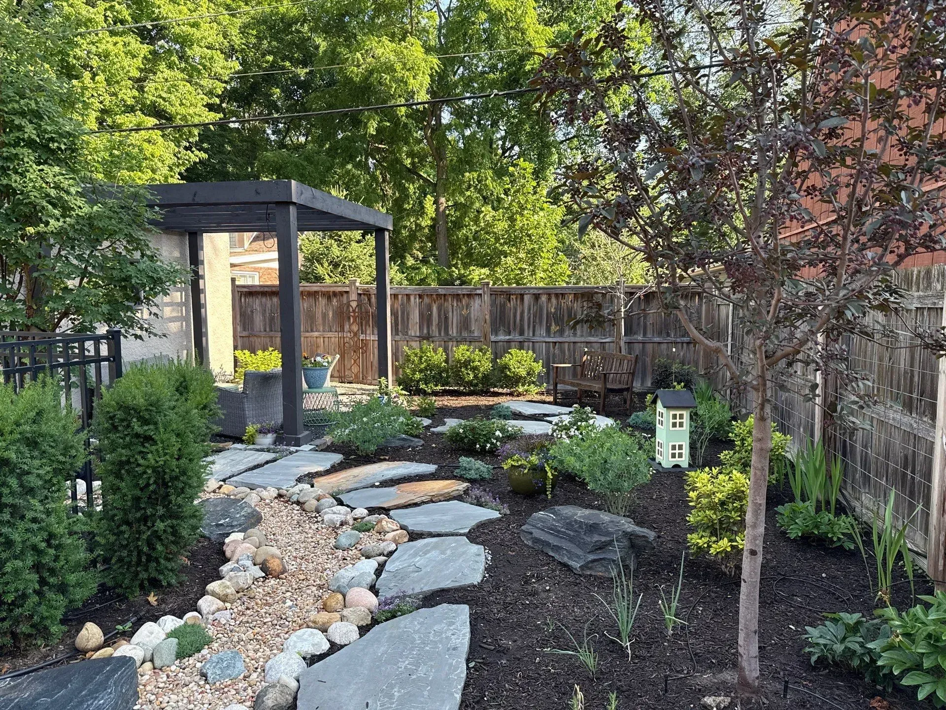 Landscaped backyard with stone path, dark mulch, trees, fence, and small arbor under a canopy of greenery