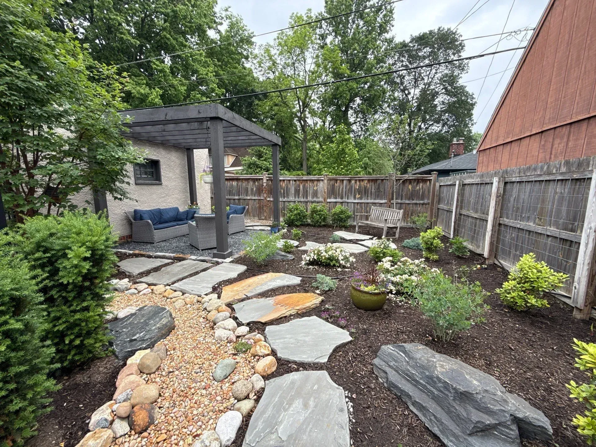 Backyard garden with stone path, gravel beds, shrubs, and a small shed beside a wooden fence.