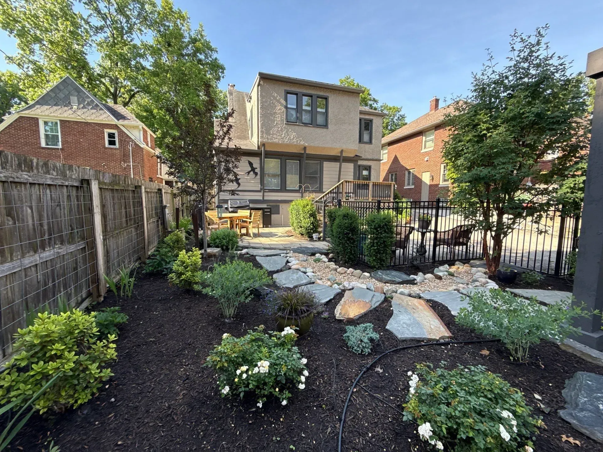 Landscaped backyard with stone path, shrubs, and a two-story house under a clear sky