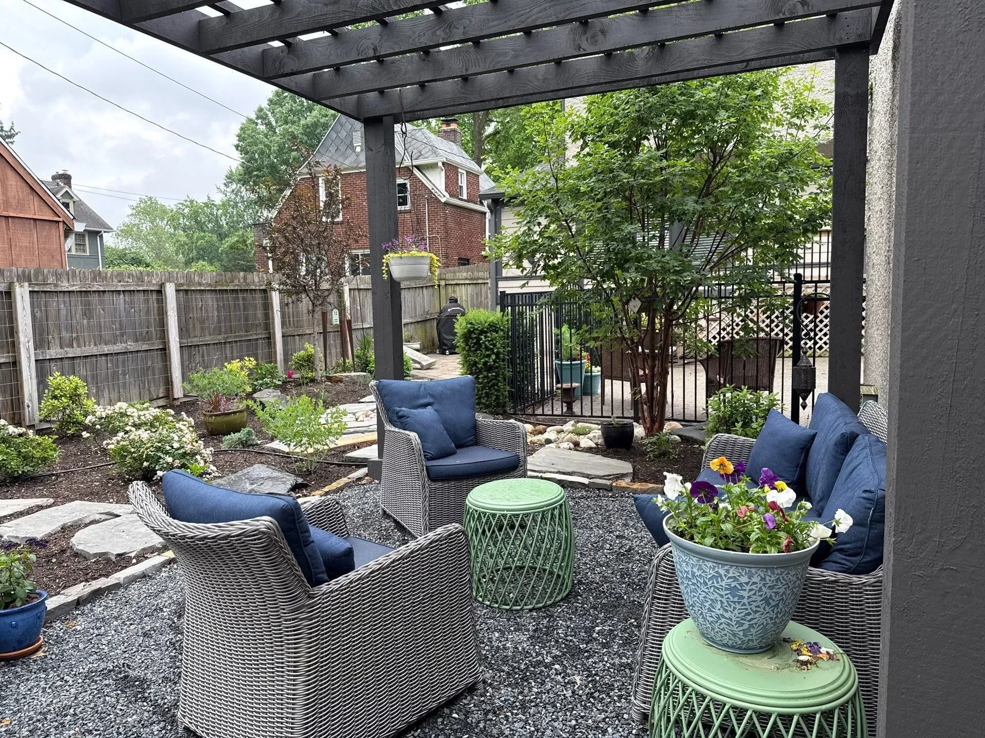 Cozy patio with blue cushioned wicker chairs, potted flowers, and a pergola beside a fenced garden