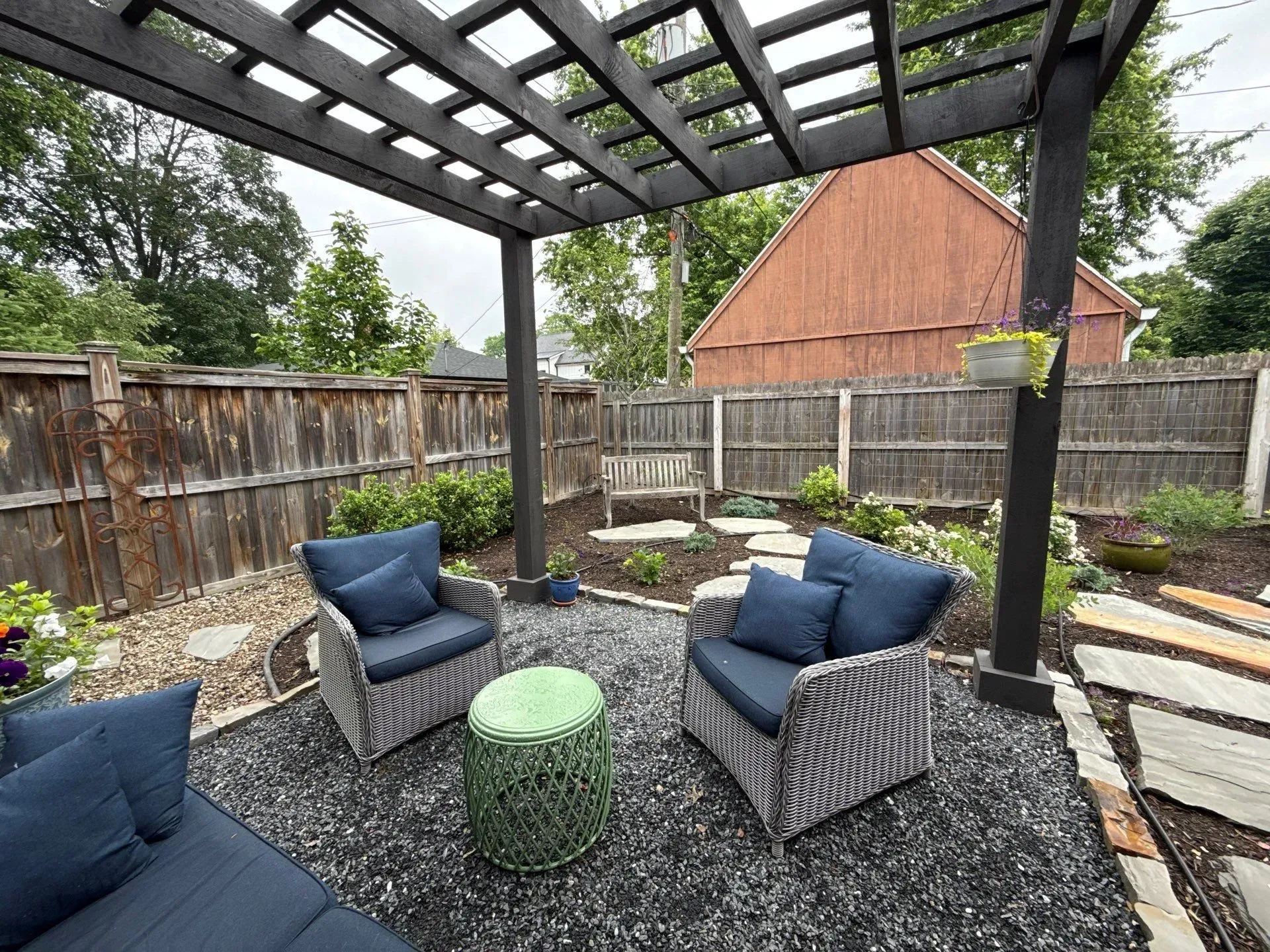 Backyard patio with black pergola, navy chairs, green ottoman, gravel ground, and fenced garden area
