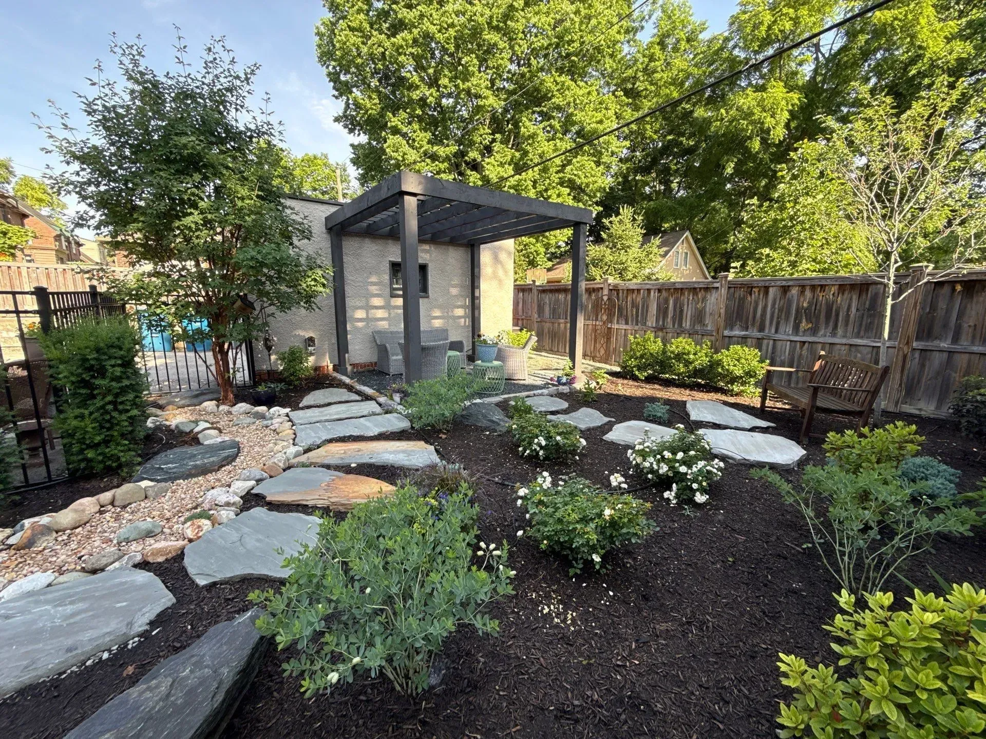 Backyard garden with stone path, dark mulch beds, small shrubs, and a gray shed with pergola overhang