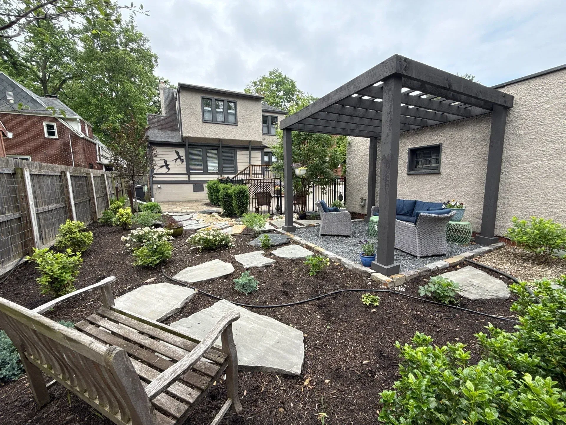 Backyard patio with stone paths, pergola, shrubs, and wooden bench beside a house