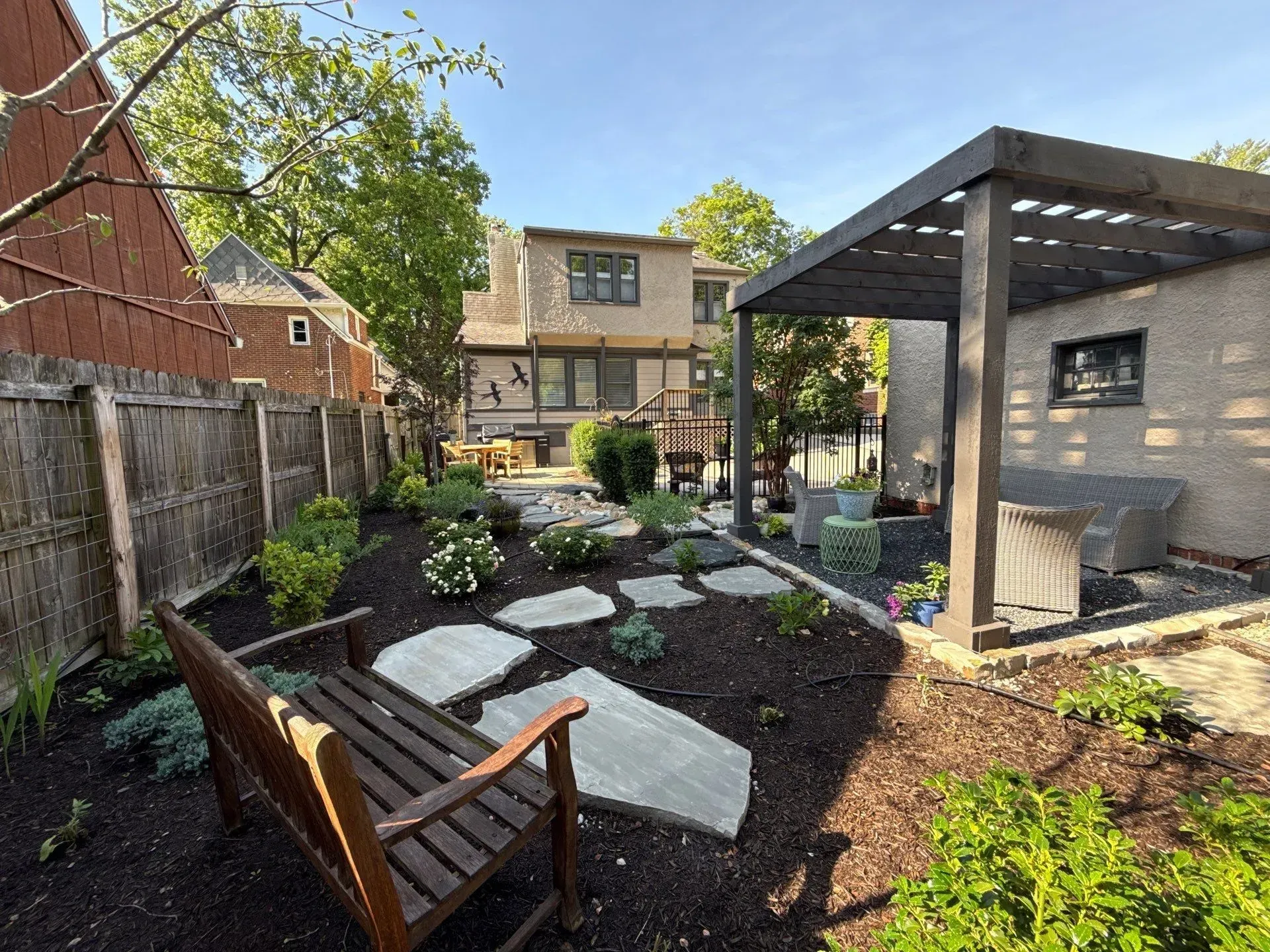 Small landscaped backyard with stone path, wooden chairs, and a modern pergola beside a house.
