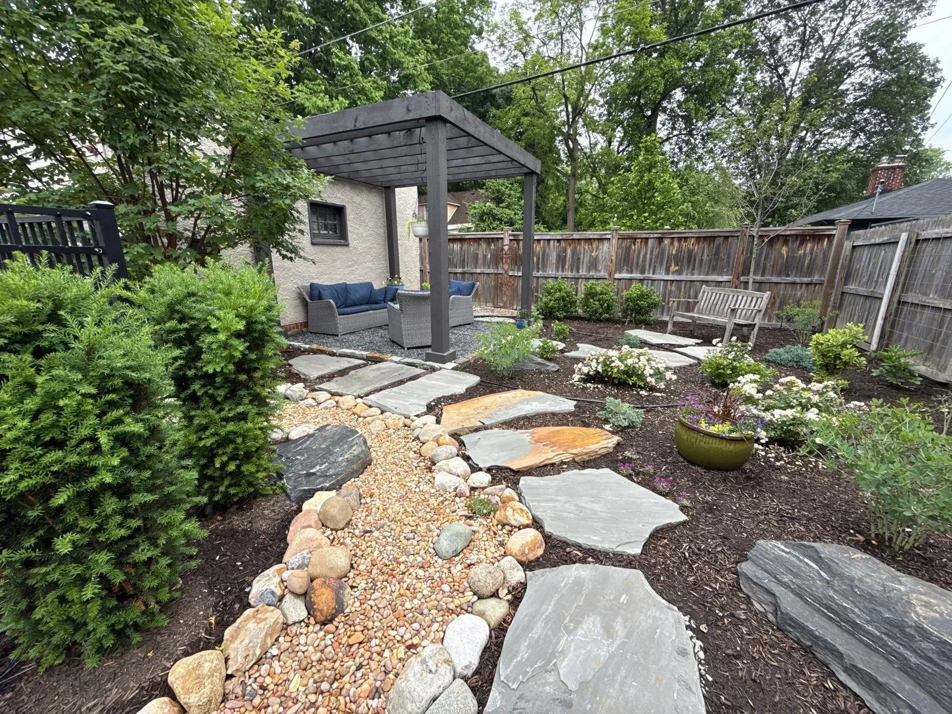 Backyard garden with stone path, pebble bed, gazebo, shrubs, and wooden fence