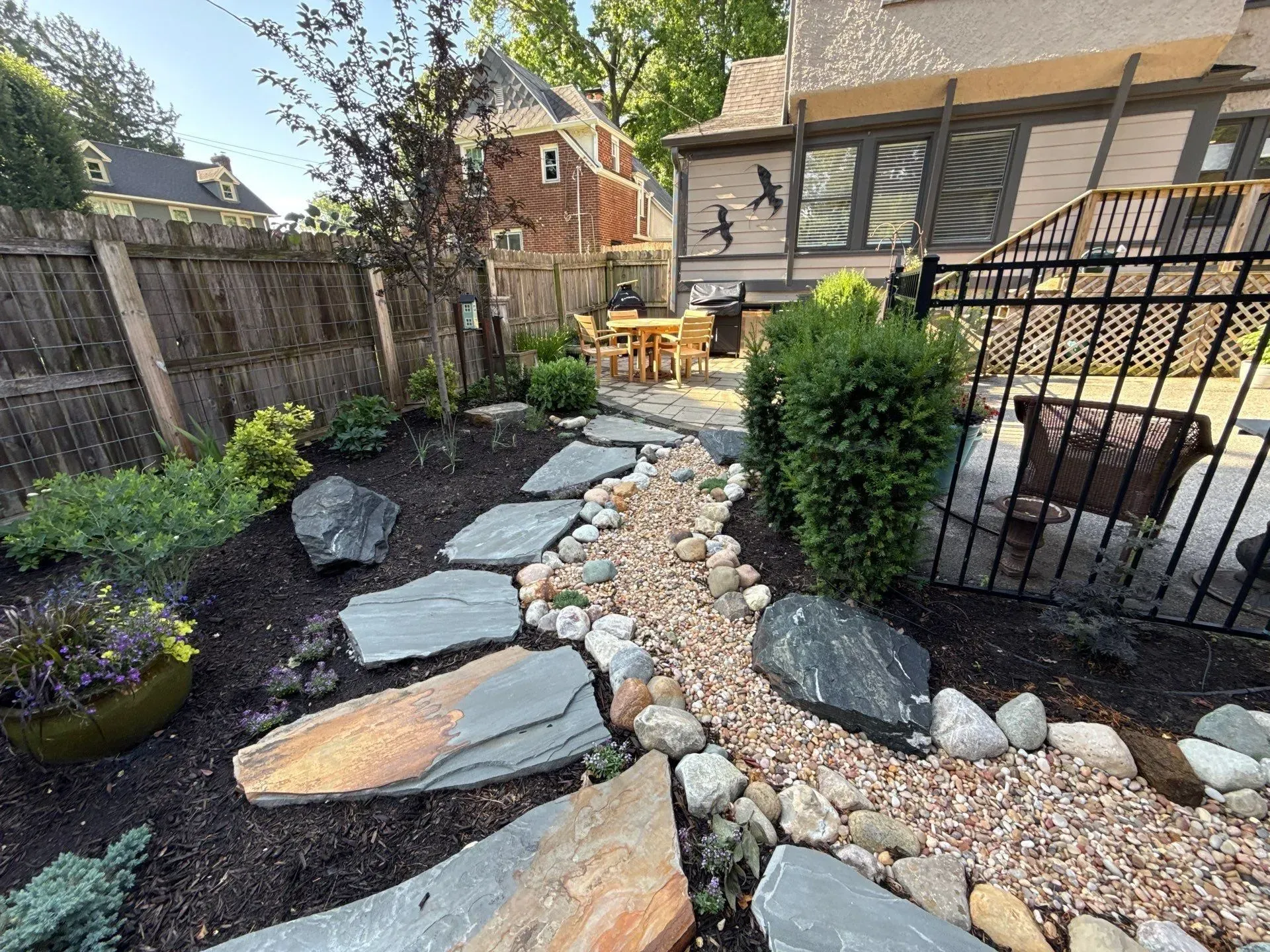 Stone garden path with slate stepping stones, pebbles, and shrubs beside a fenced backyard patio.