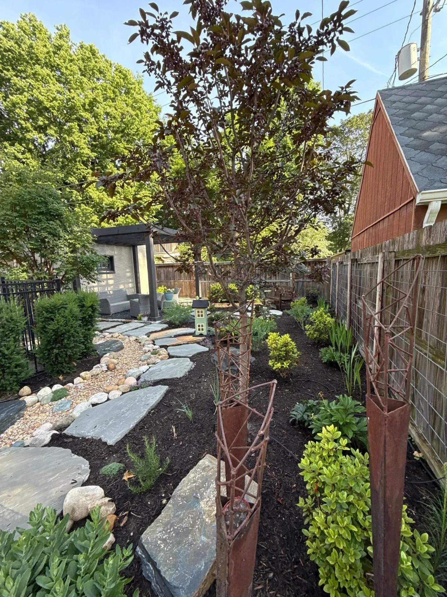 Narrow landscaped garden path with flagstones, dark mulch, shrubs, and young trees beside a wooden fence.