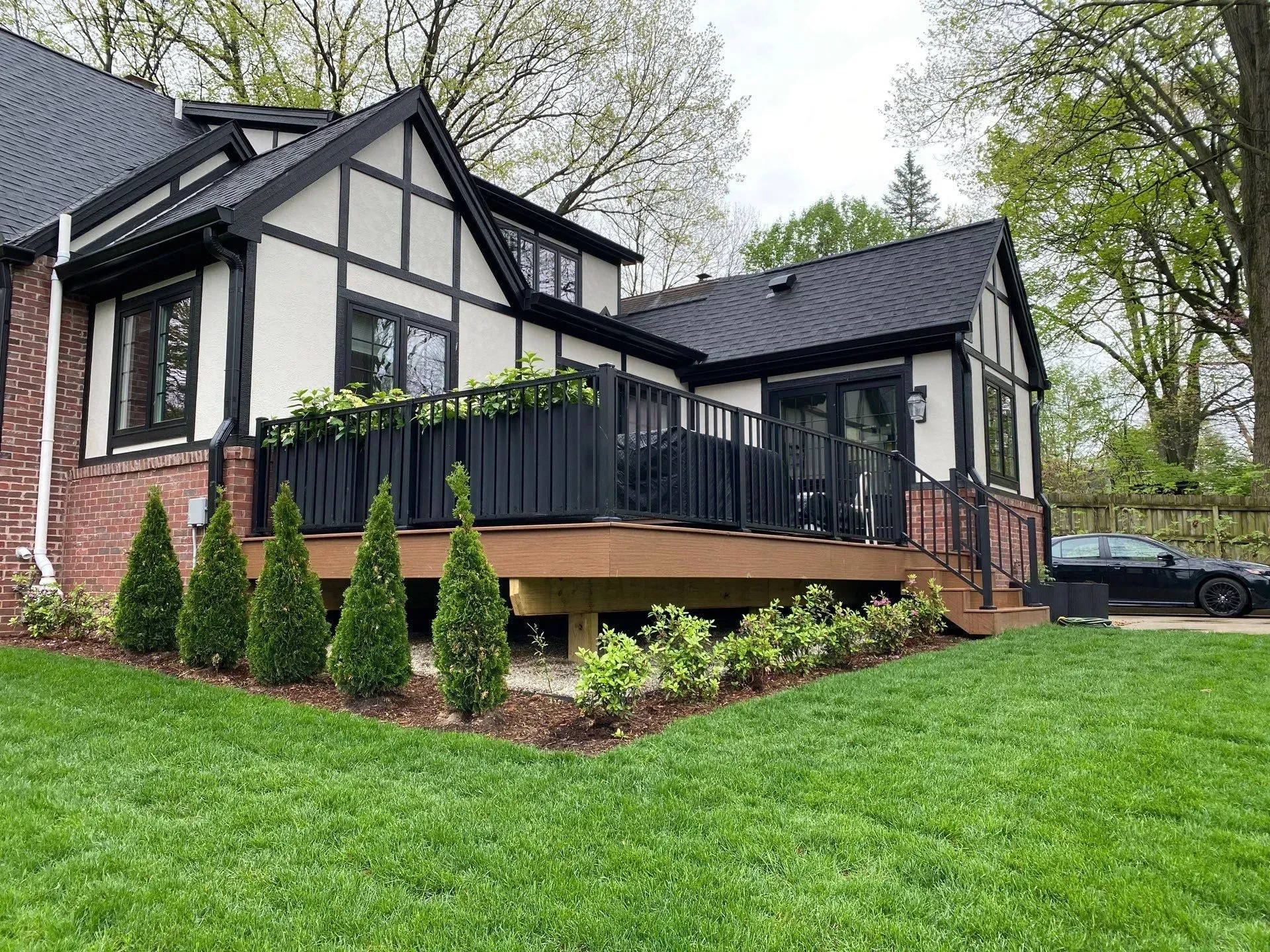 Black-and-white Tudor-style house with a wooden deck, black railings, and green lawn in a wooded yard