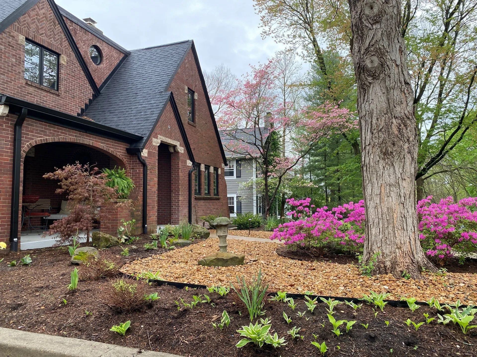 Brick house with a lush garden, blooming pink flowers, and a large tree in springtime