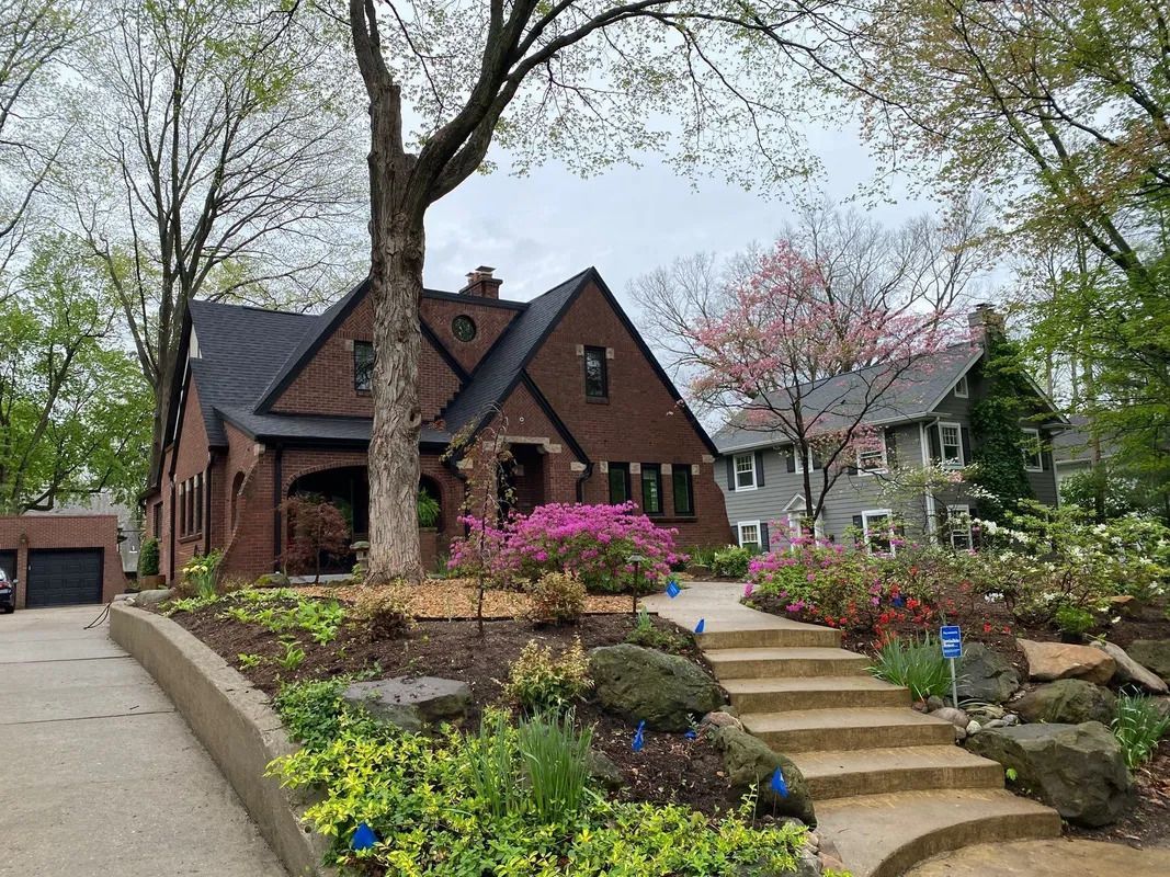 Brick house with steep roof, blooming garden, and stone steps under tall trees