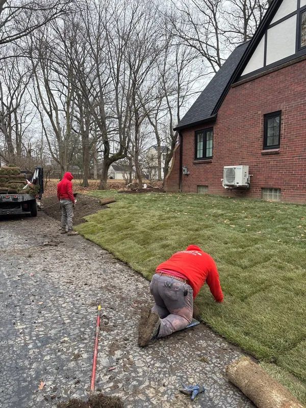 Two workers in red jackets installing grass beside a brick house on a gravel driveway.