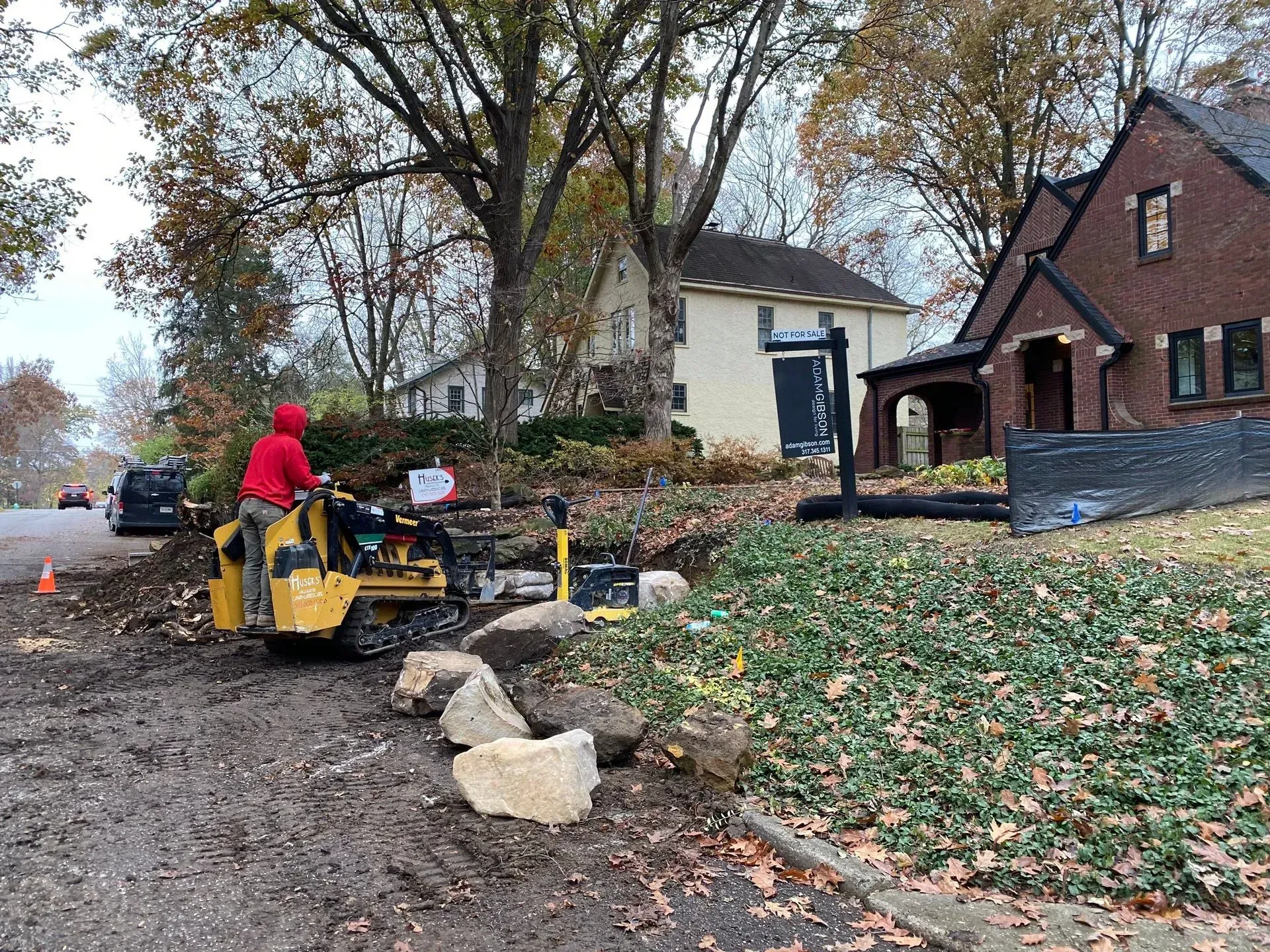 Workers use a small bulldozer to clear leaves along a muddy suburban street beside houses and trees