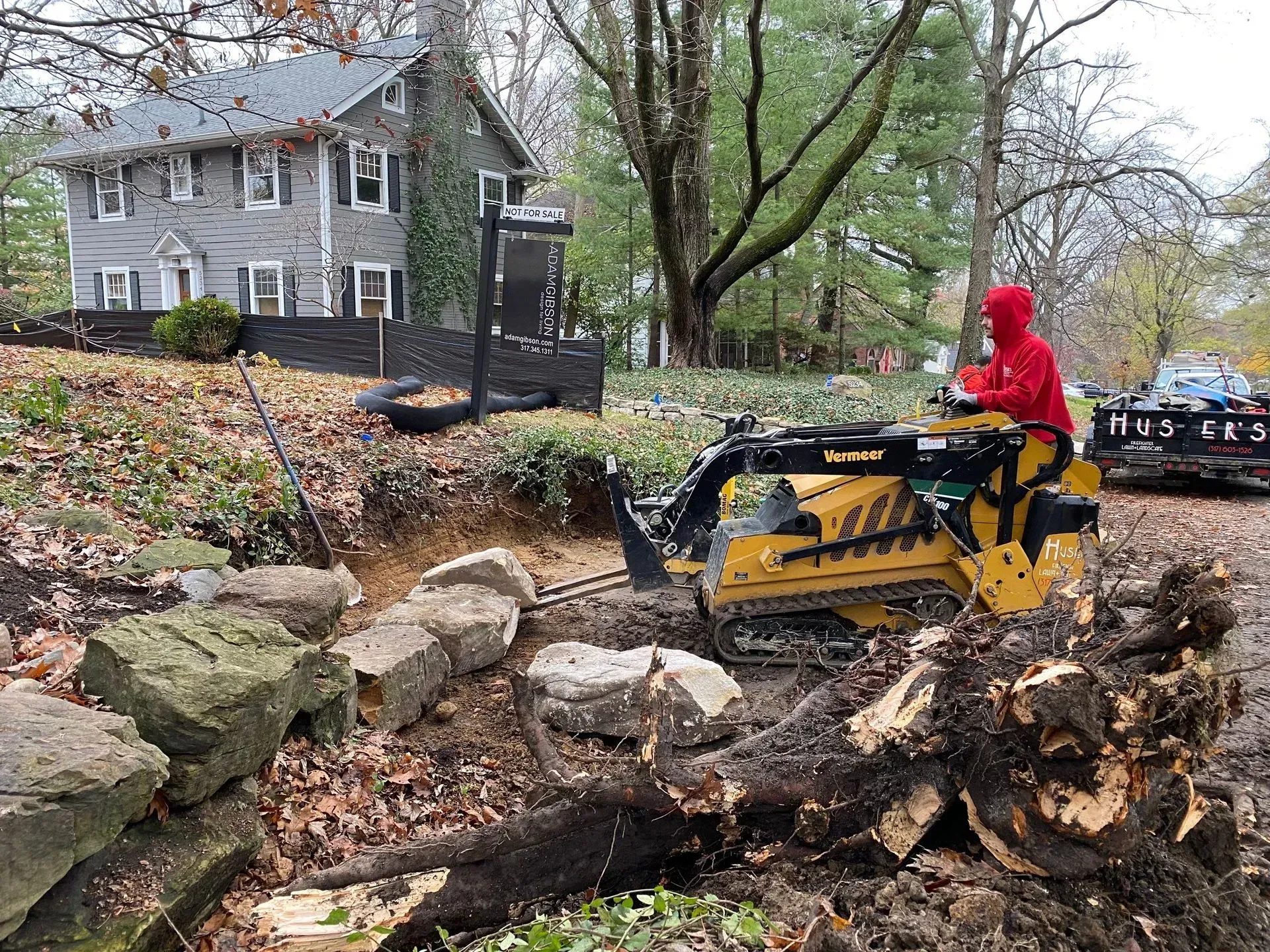 Person in red hoodie operating a yellow mini excavator clearing debris beside a rocky yard and house