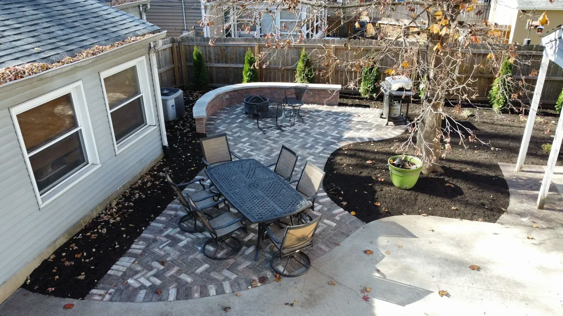 Patio with round stone table, black chairs, potted plant, and curved brick wall beside a house