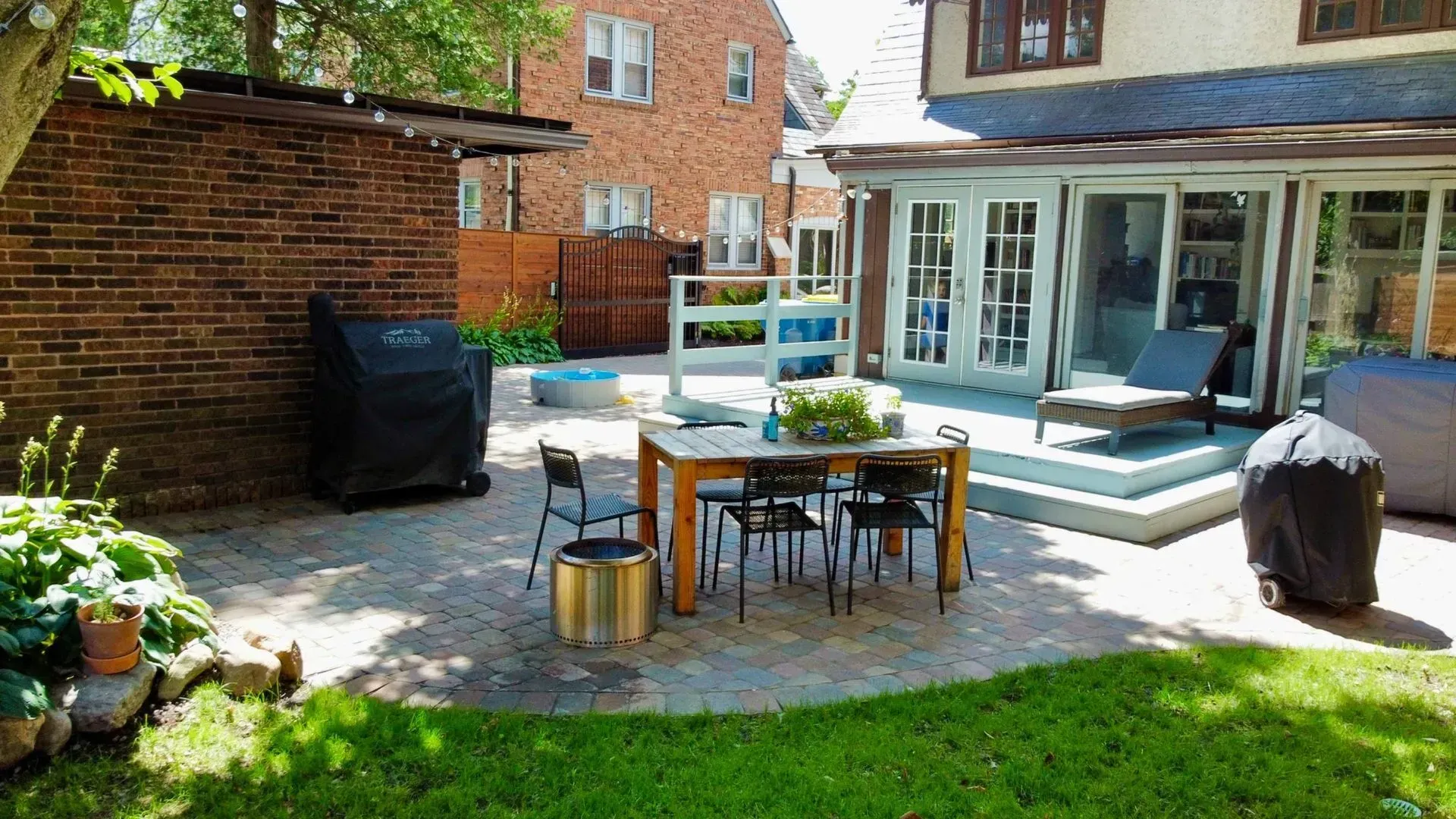 Patio with dining table, chairs, lounge chairs, grill, and potted plants in a sunny backyard