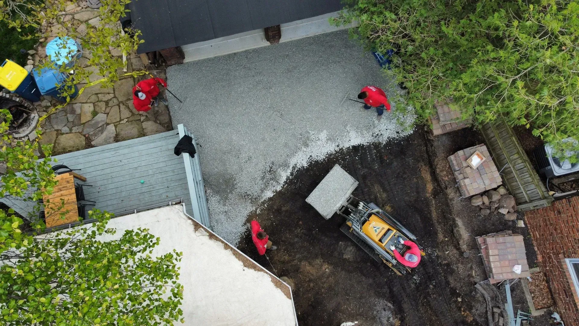 Aerial view of workers in red gear around a backhoe on a dirt lot beside a driveway and trees