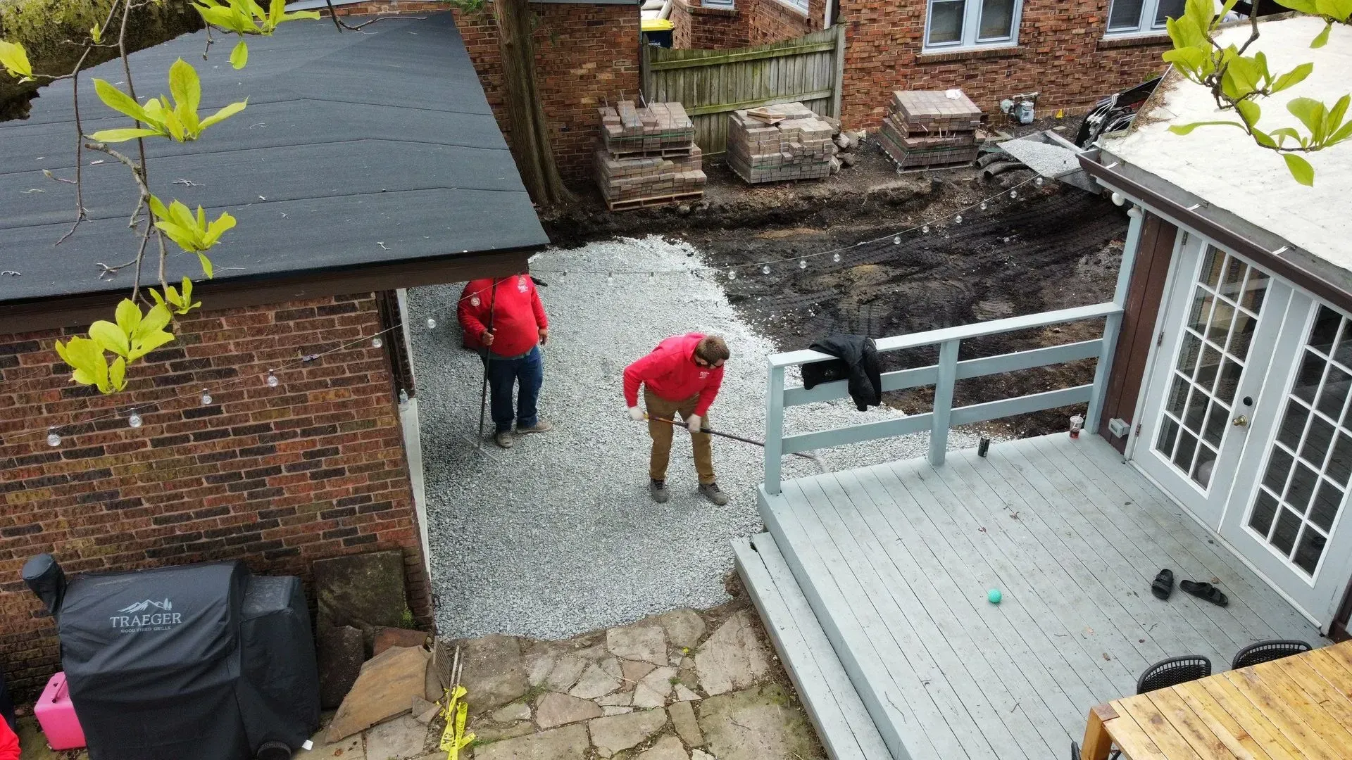 Two people in red jackets work in a gravel courtyard beside a brick house and white deck.