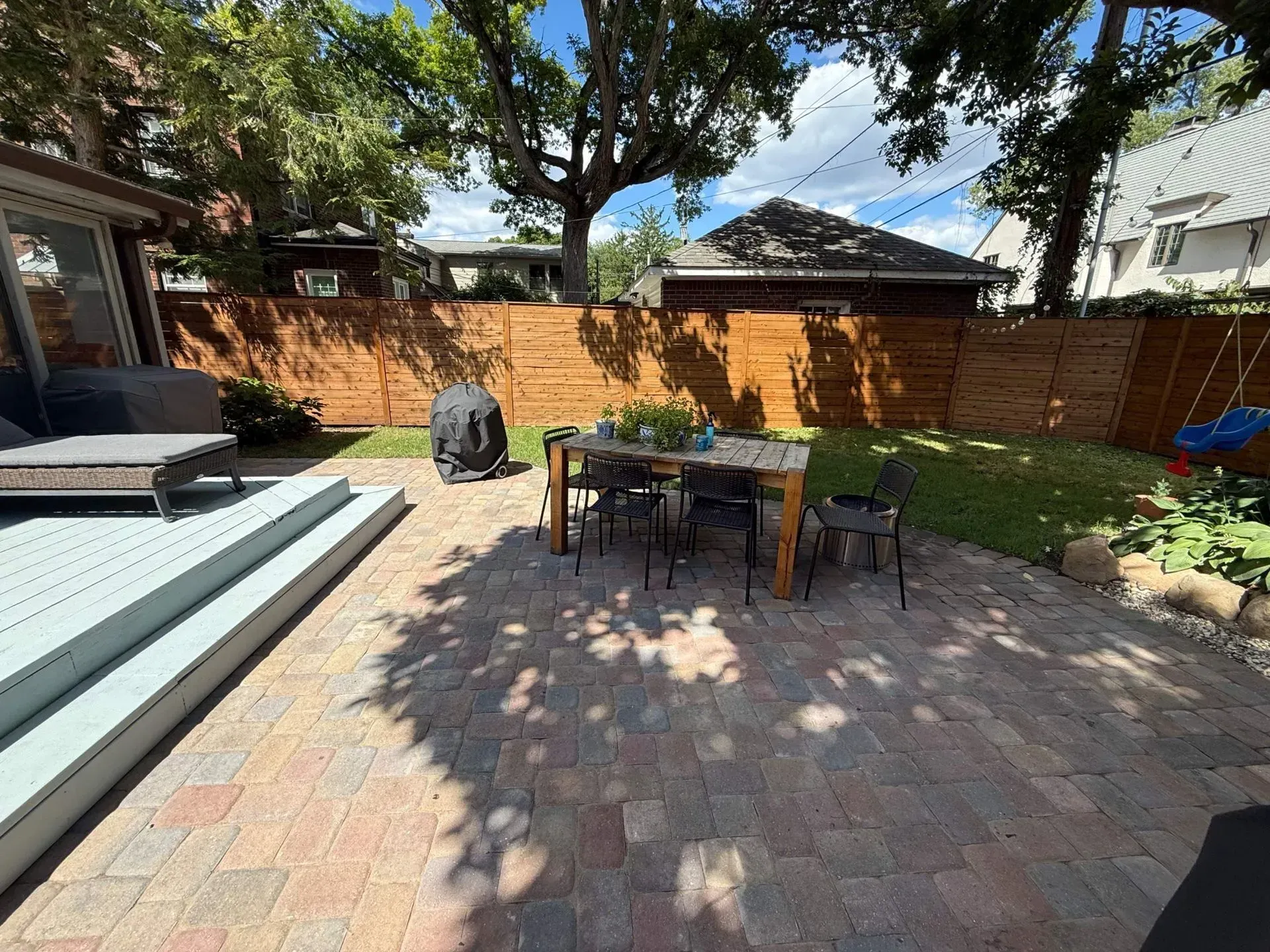 Backyard patio with table, chairs, grill, and paver seating area under trees and a wooden fence.