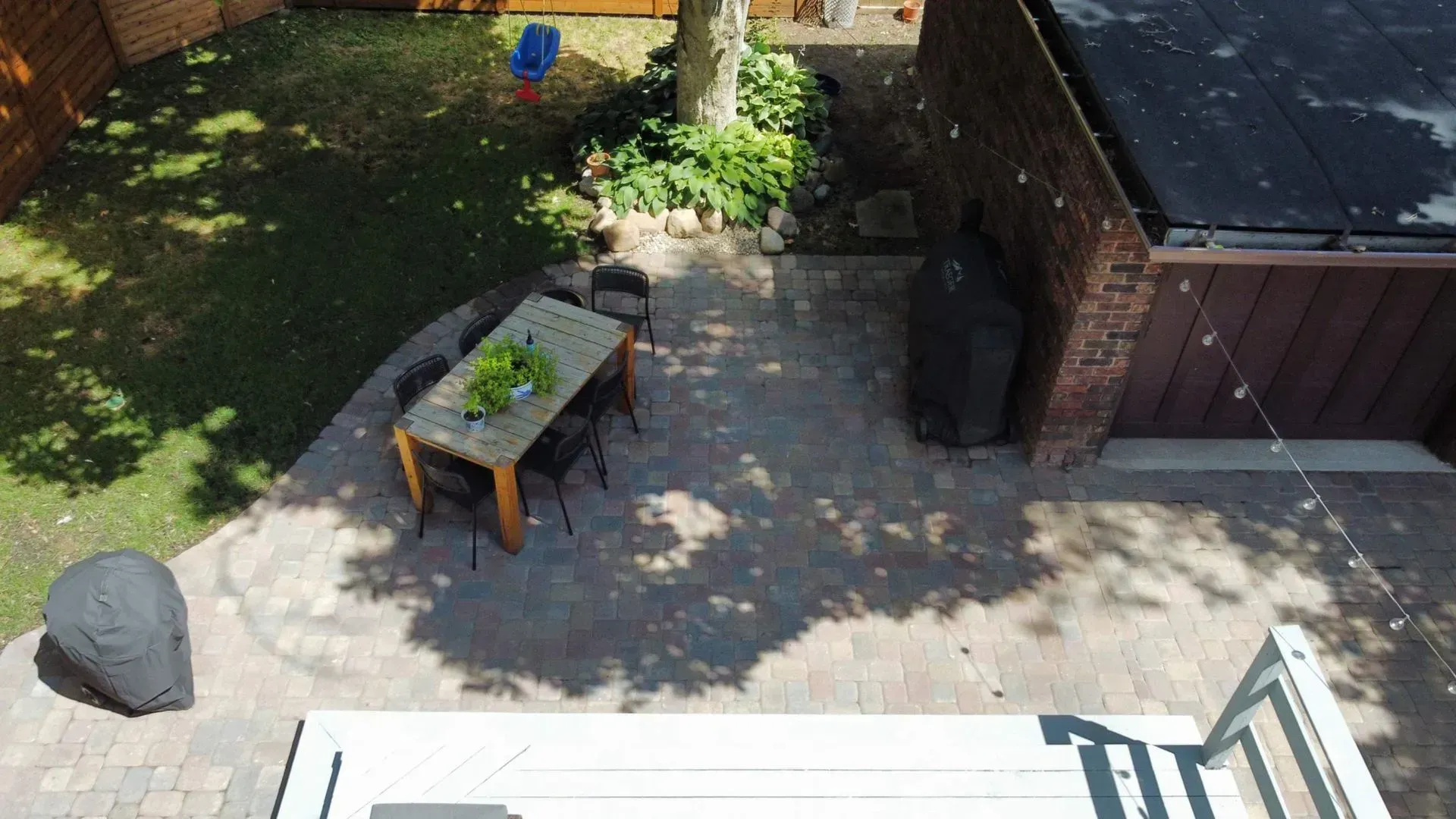 Backyard patio with table, chairs, grill, shed, and a small lawn in sunlight.