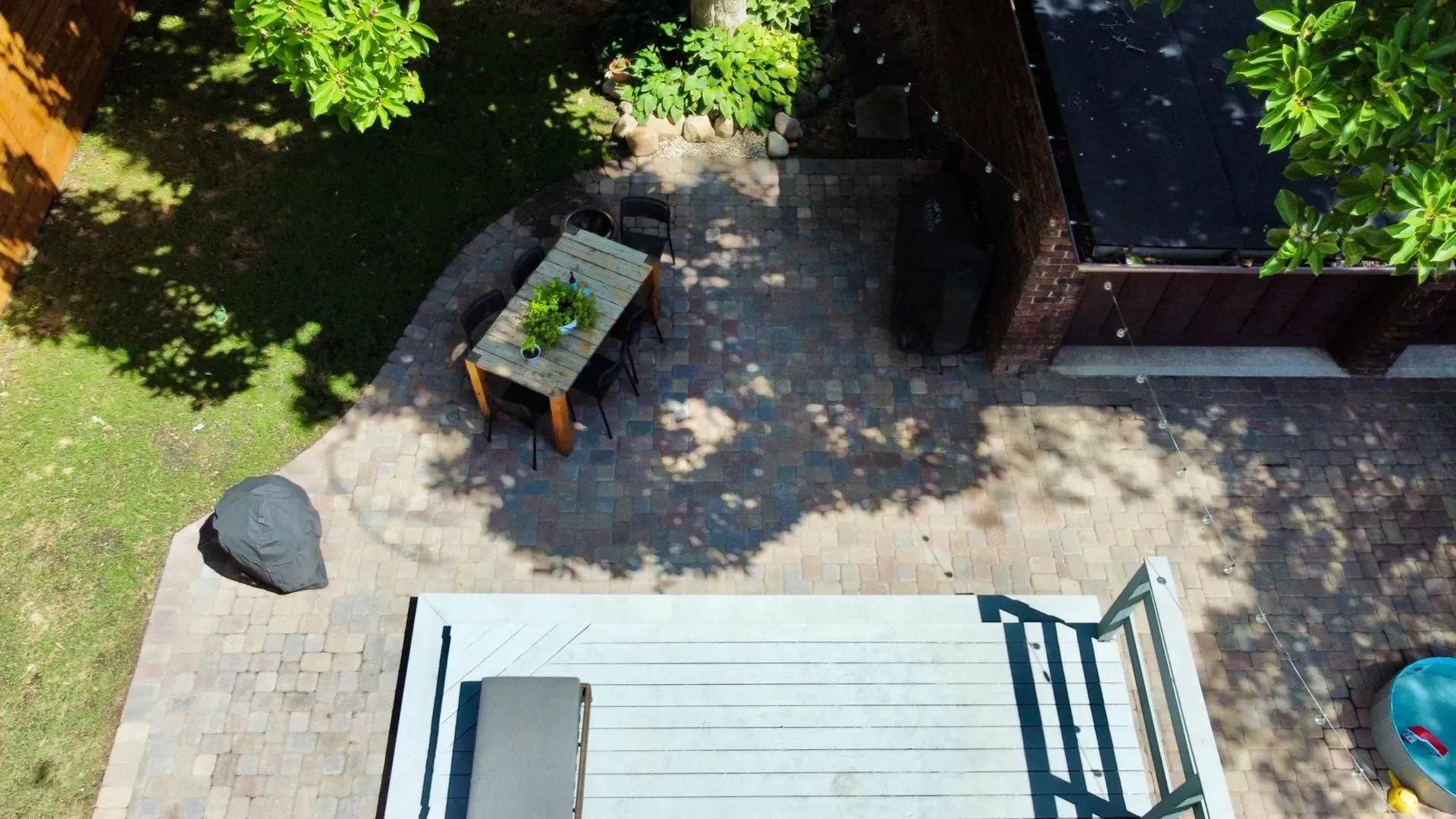 Backyard patio with a table and chair, paving stones, and a stair railing beside greenery and a shed.
