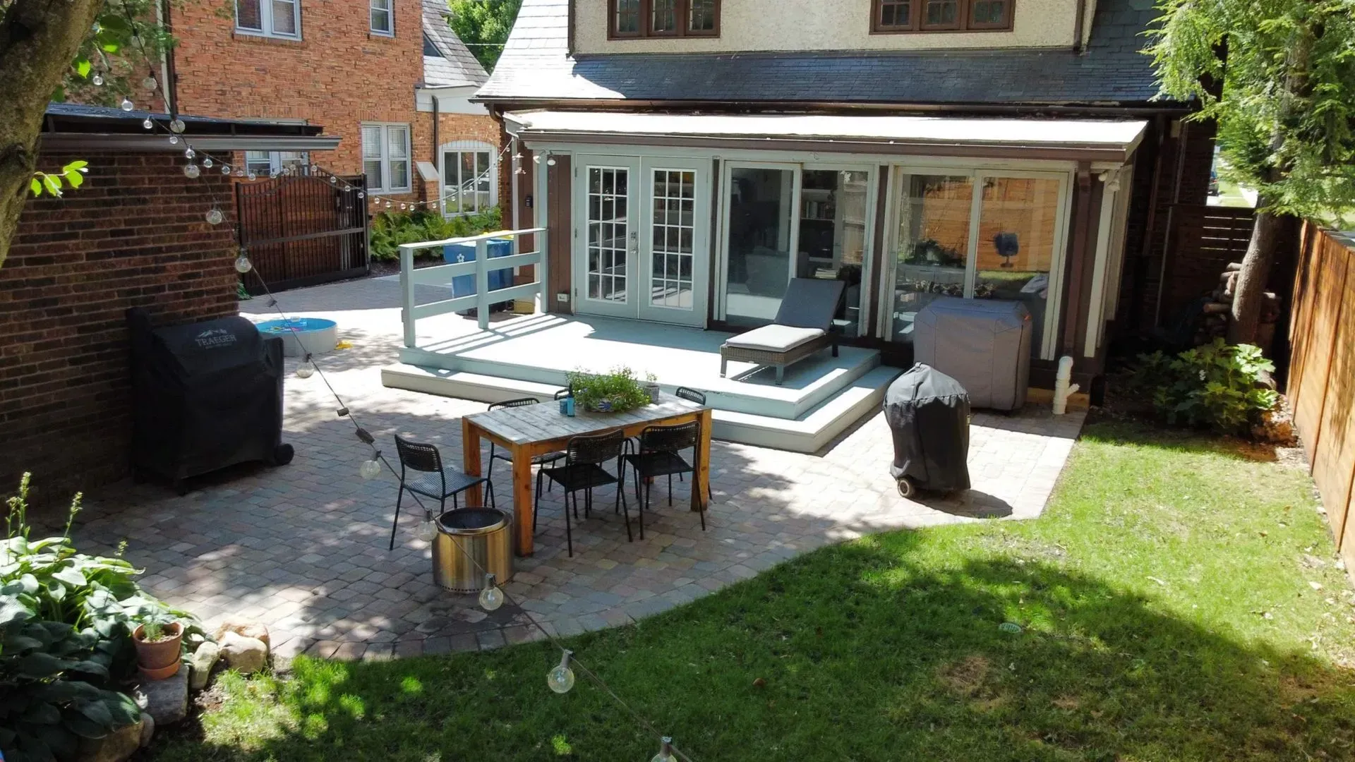 Backyard patio with outdoor dining table, gray seating, and a glass-walled house opening onto a lawn
