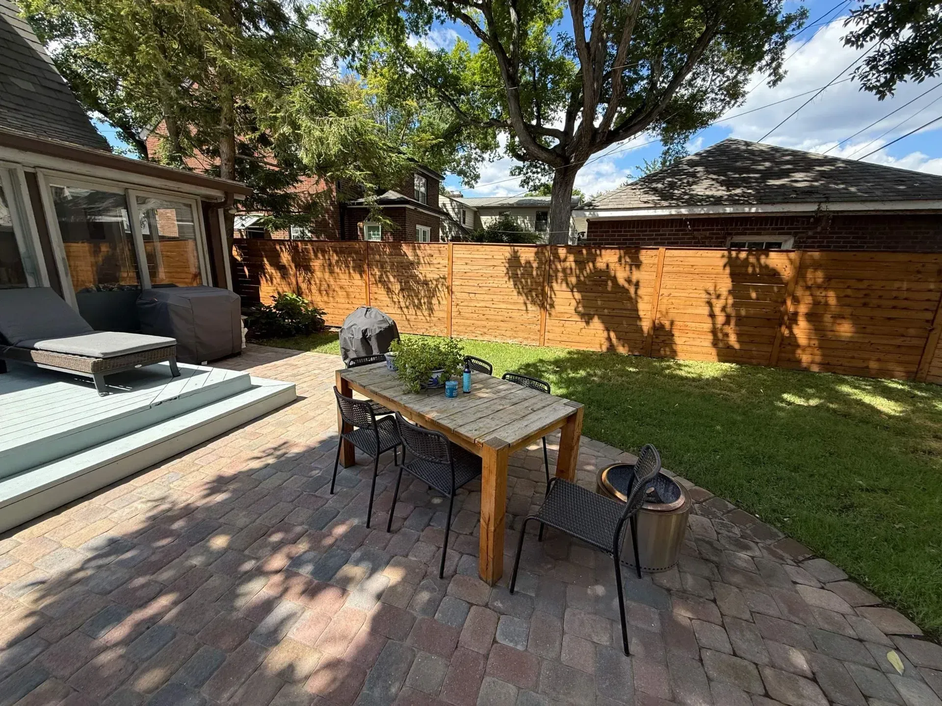 Patio with outdoor table and chairs, grill, brick pavers, and wooden fence in a sunny backyard