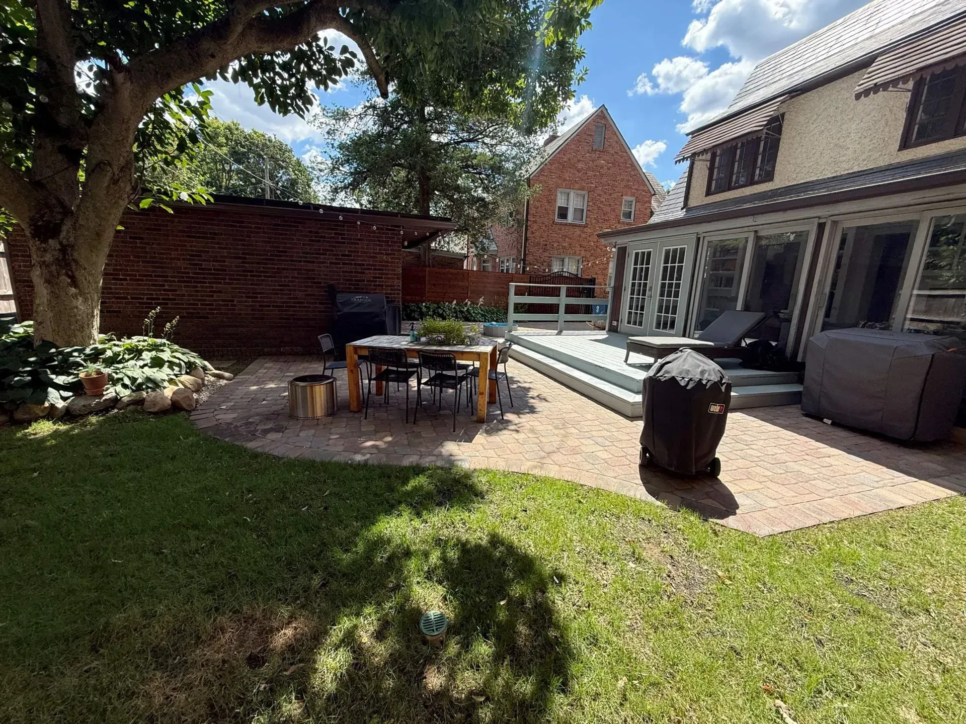 Backyard patio with outdoor seating, lawn, trees, and a brick house with large windows