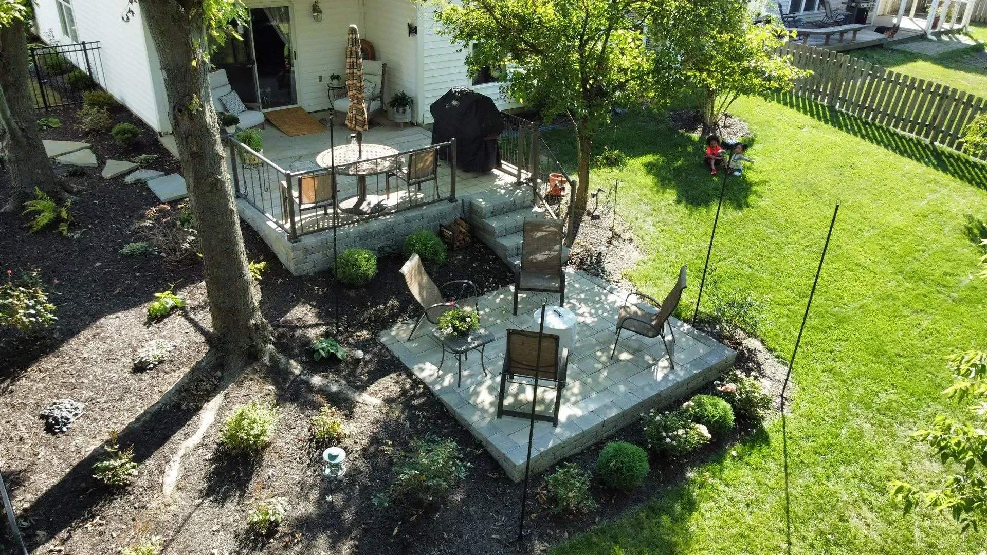 Backyard patio with two stone seating areas, chairs, and a grill beside a white house and green lawn