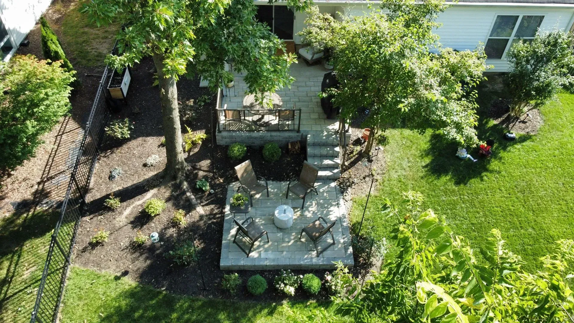 Backyard patio with stone table and chairs under trees beside a green lawn